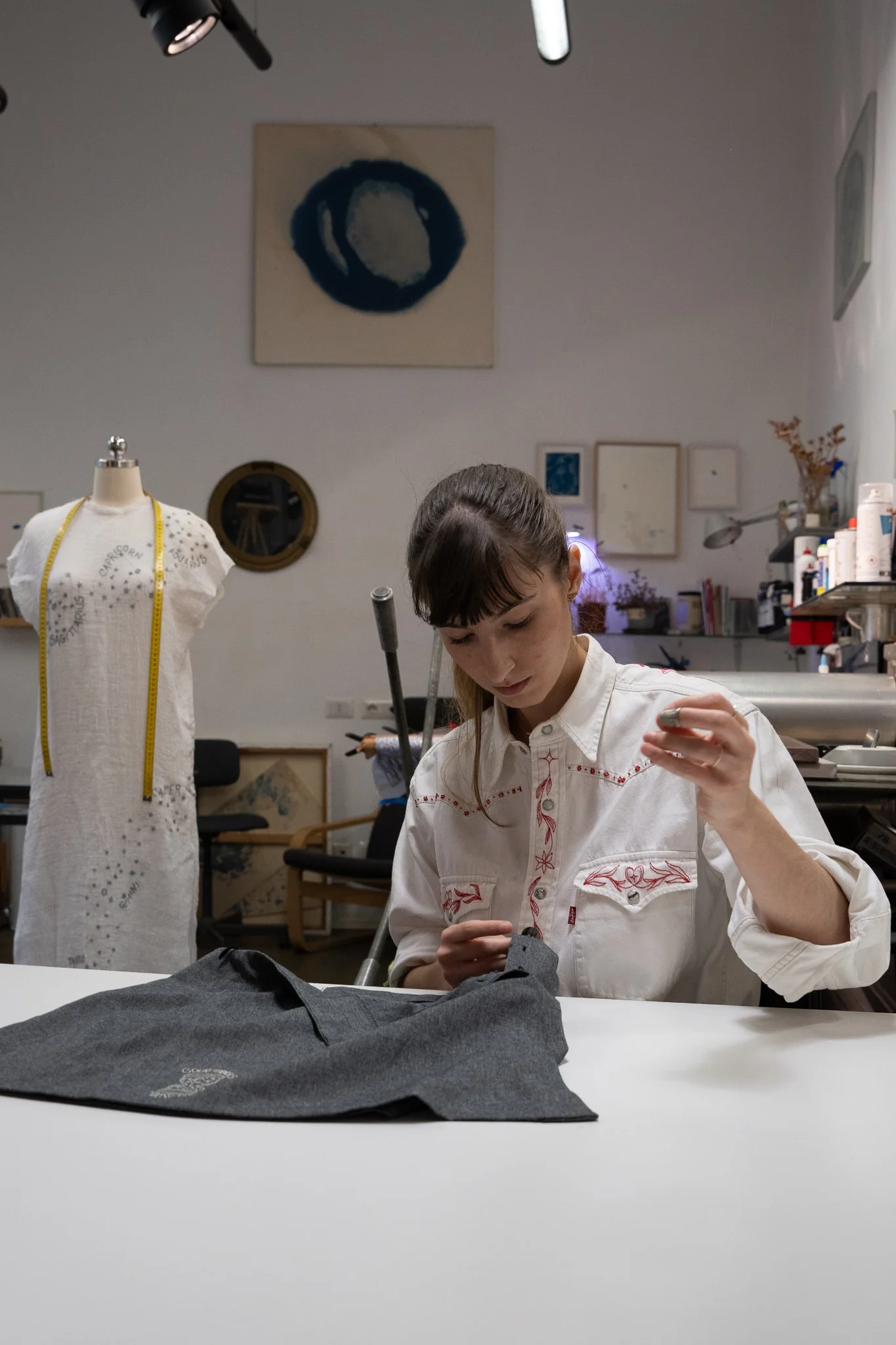 A woman working on sewing or tailoring at a craft table in a creative studio decorated with artworks, fabrics, and tools.