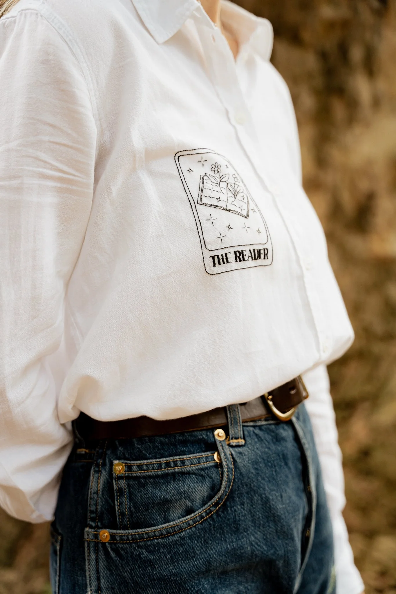 Close-up of a person wearing a white shirt with a graphic of a book, flowers, and stars on it, and blue jeans with a belt, outdoors.