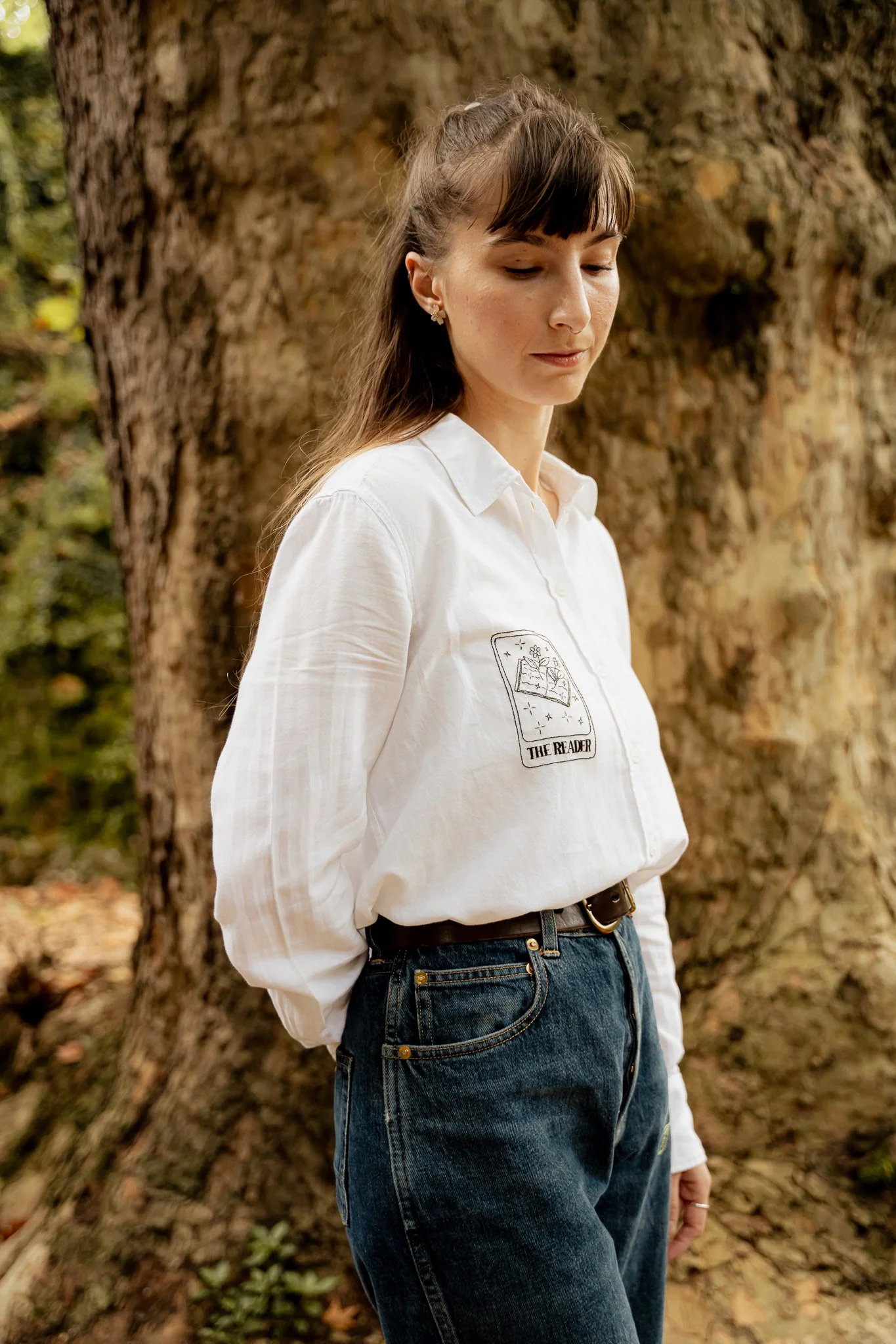 A young woman stands in front of a large tree trunk, wearing a white button-up shirt with an embroiderer tarot card on the chest and dark blue jeans.