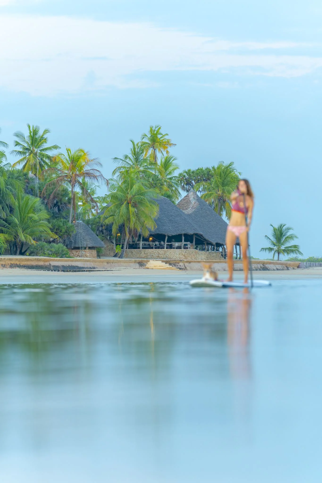 A woman standing on a paddleboard in the water near a tropical beach with palm trees and thatched roof huts in the background.