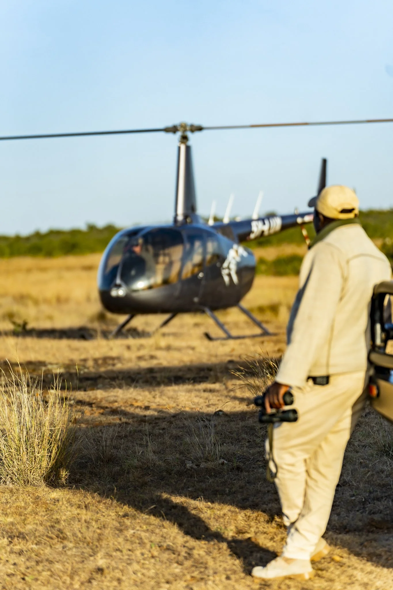 A person dressed in light-colored clothing and a yellow cap walking with a remote control in hand in front of a black helicopter on a grassy field.