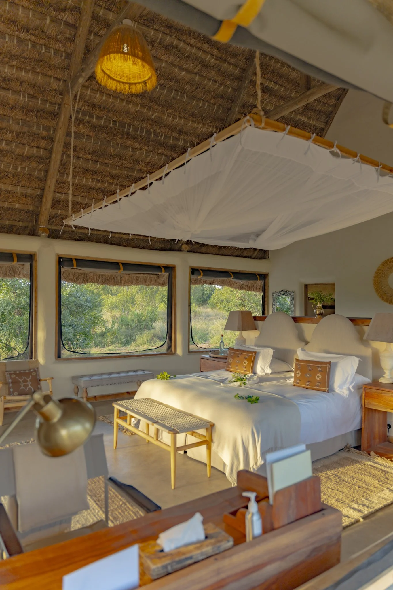 A cozy bedroom with a large bed covered in white linens, decorated with small yellow pillows and green foliage accents. The room has a thatched roof and large windows showing greenery outside. There is a wooden side table with a lamp and a bottle on 