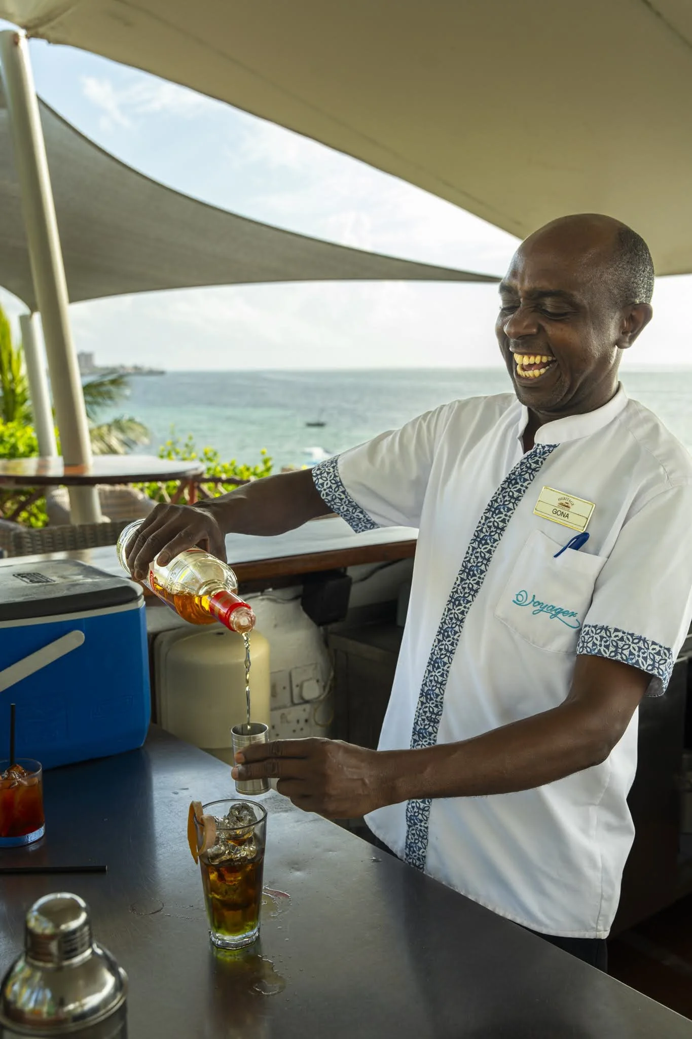 A smiling man wearing a white shirt with blue patterns on the collar and sleeves is pouring a drink into a glass with ice at an outdoor bar, with the ocean and sky in the background.