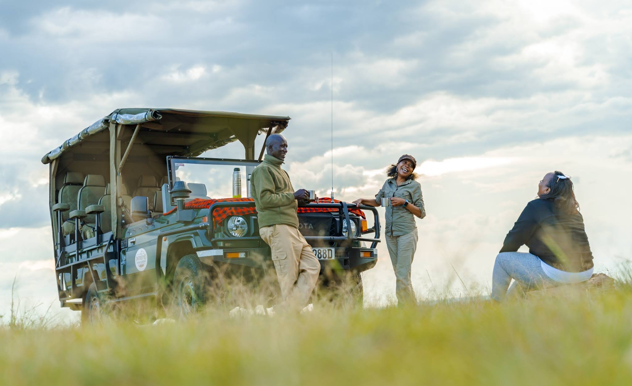 Three people outdoors near a safari vehicle with an open roof, talking and smiling under a cloudy sky.