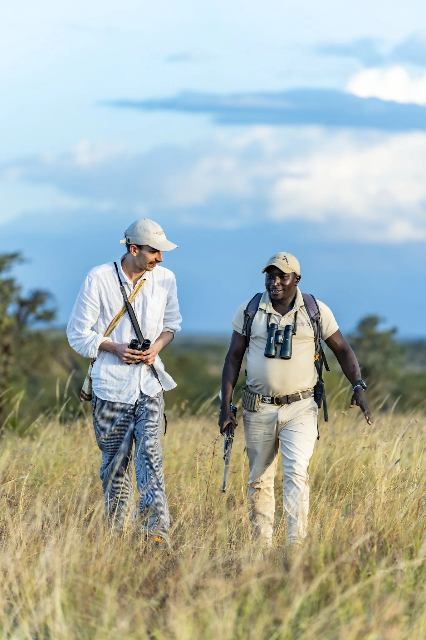Two men walk through a grassy field with binoculars around their necks, talking to each other. One man is wearing a white shirt and plaid pants, and the other is wearing a beige shirt and light-colored pants. They are both wearing hats.