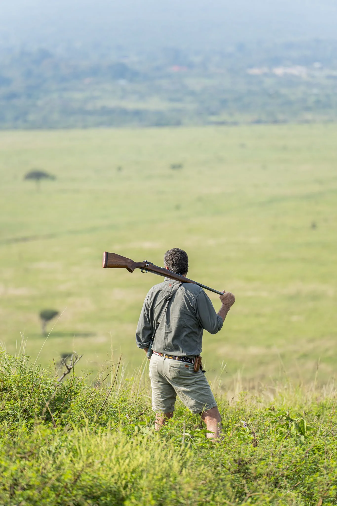 A person standing in a grassy field, holding a rifle over their shoulder, facing away from the camera, with hills and trees in the background.