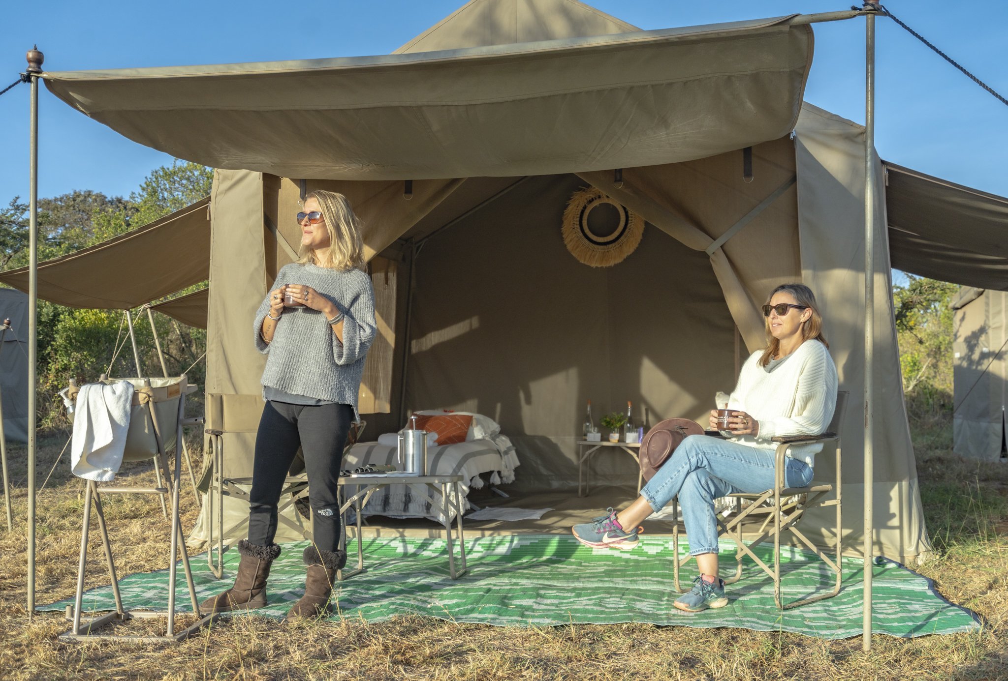Two women are relaxing outside a large beige camping tent on a green checkered mat. One woman is standing, wearing sunglasses, a gray sweater, black pants, and brown boots, holding a drink. The other woman is sitting in a folding chair, also with sunglasses, wearing a white sweater, jeans, and sneakers, holding a drink. The tent has an open entrance, with a bed and some items visible inside. The background features trees, a clear blue sky, and sunny weather.