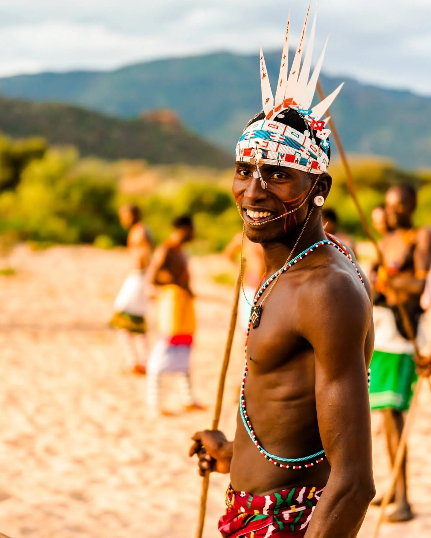 A smiling man from an indigenous African tribe wearing traditional attire and headgear with feathers, standing outdoors with mountains and blurred people in the background.