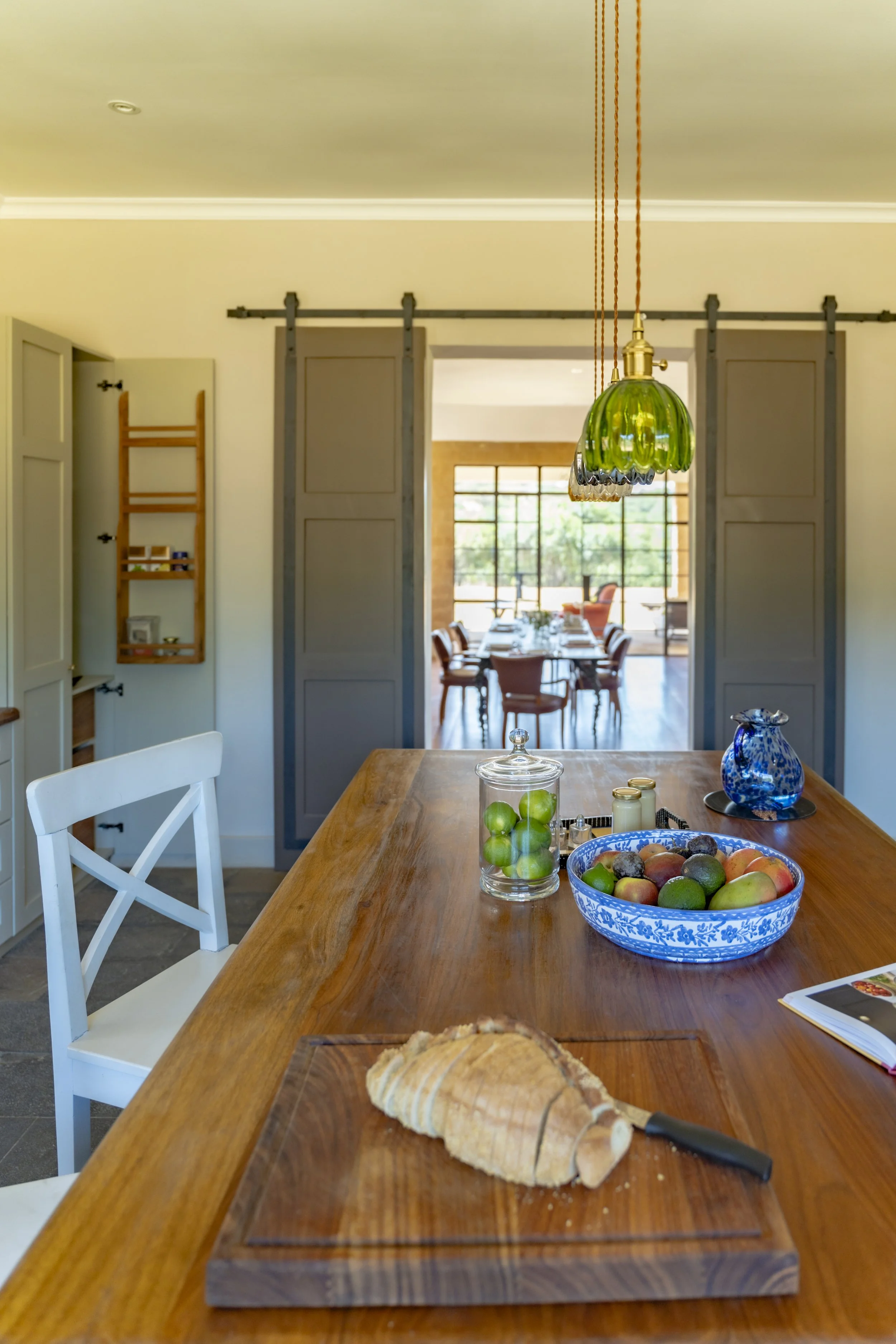 A modern dining room with a wooden table, a loaf of bread on a cutting board, a bowl of assorted fruits, a glass jar of green apples, and a blue and white pitcher. The background features a dining area with chairs, large windows, and sliding barn doo