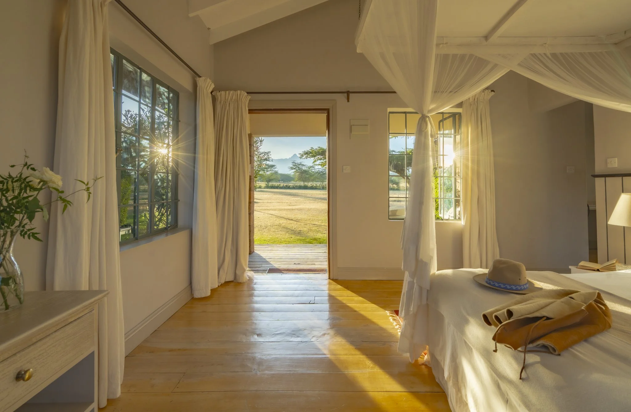 A cozy bedroom with white curtains, wooden floor, and an open door leading to a sunny outdoor landscape with trees and mountains.