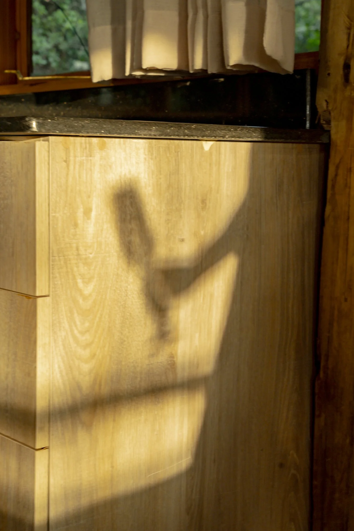 A kitchen with a wooden cabinet, a black countertop, a window with cream curtains, and a shadow of a person taking a photo.