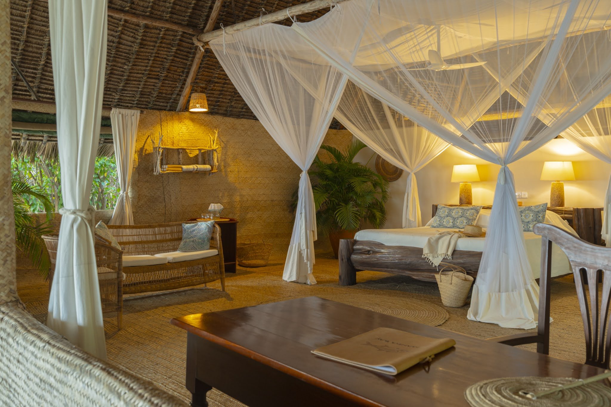 A cozy tropical bedroom with a four-poster bed draped in white mosquito netting, thatched ceiling, wooden furniture, and warm ambient lighting from lamps and a hanging wicker lampshade.