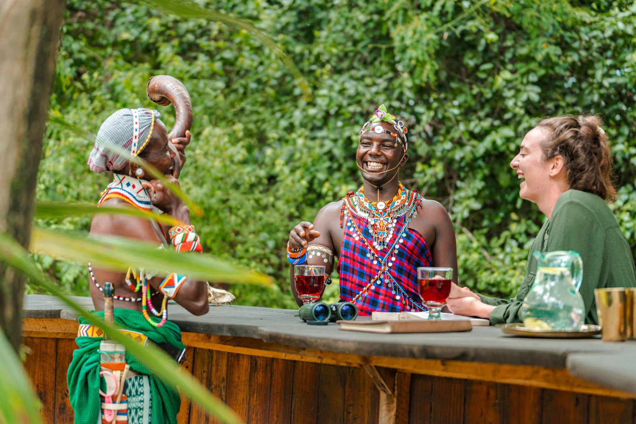 Three people dressed in traditional African attire laughing and chatting at an outdoor gathering, surrounded by green foliage.
