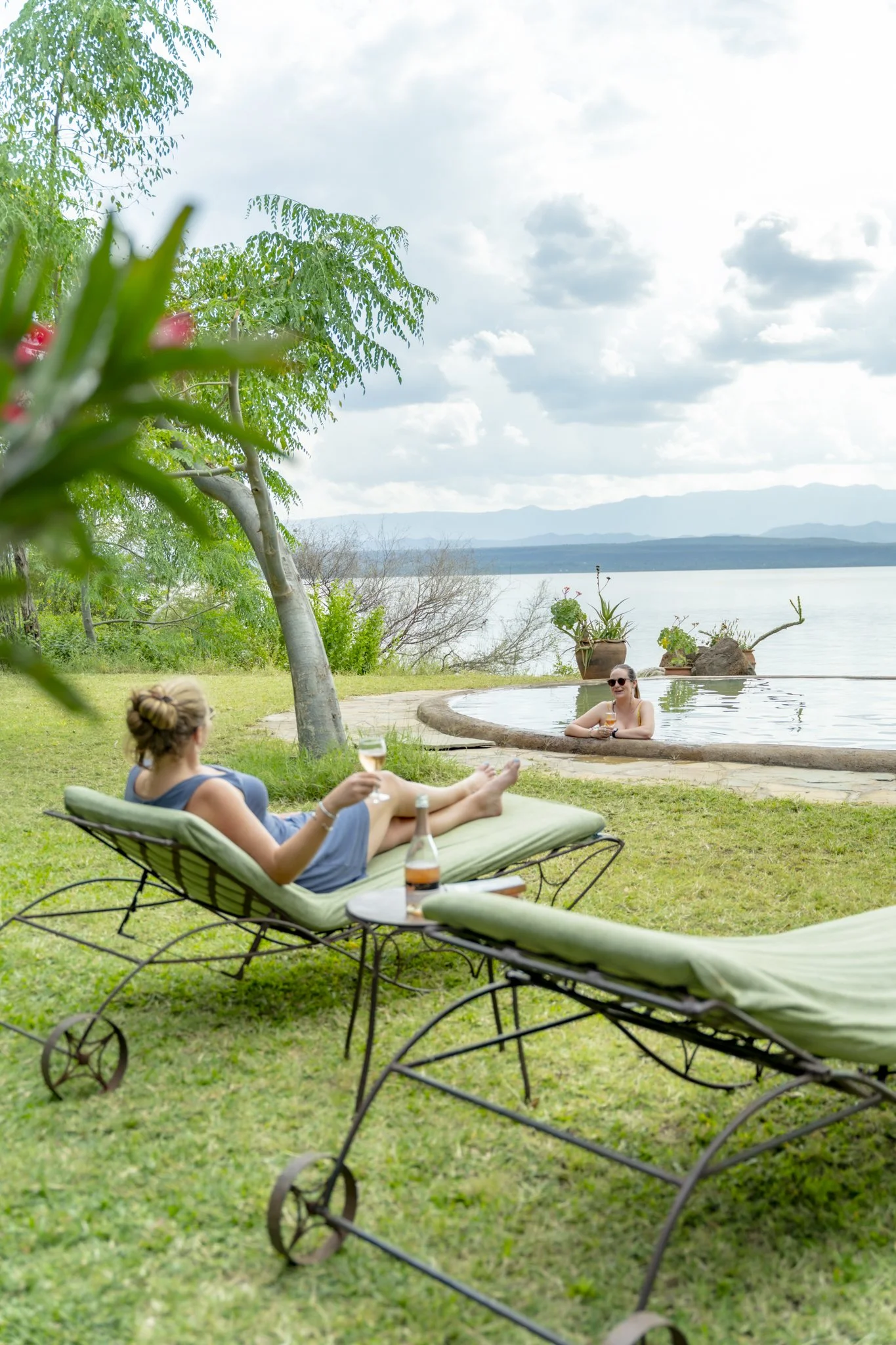 Women relaxing outdoors near a lake, sitting on lounge chairs and in a small pool, with a scenic view of mountains and a cloudy sky.