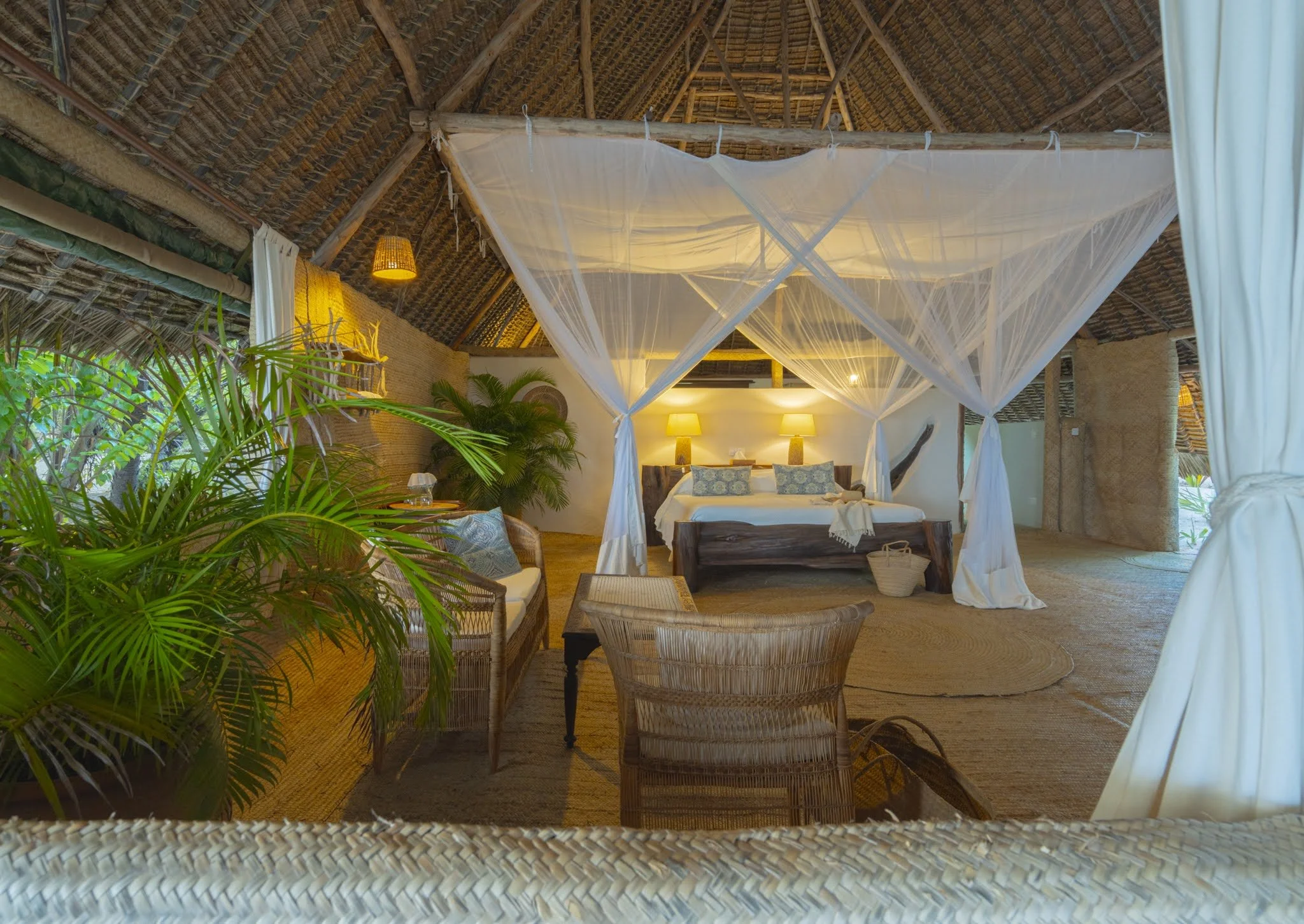 A tropical bungalow bedroom with a thatched roof, a four-poster bed with white curtains, palm trees, plants, warm lighting, woven furniture, and natural decor.