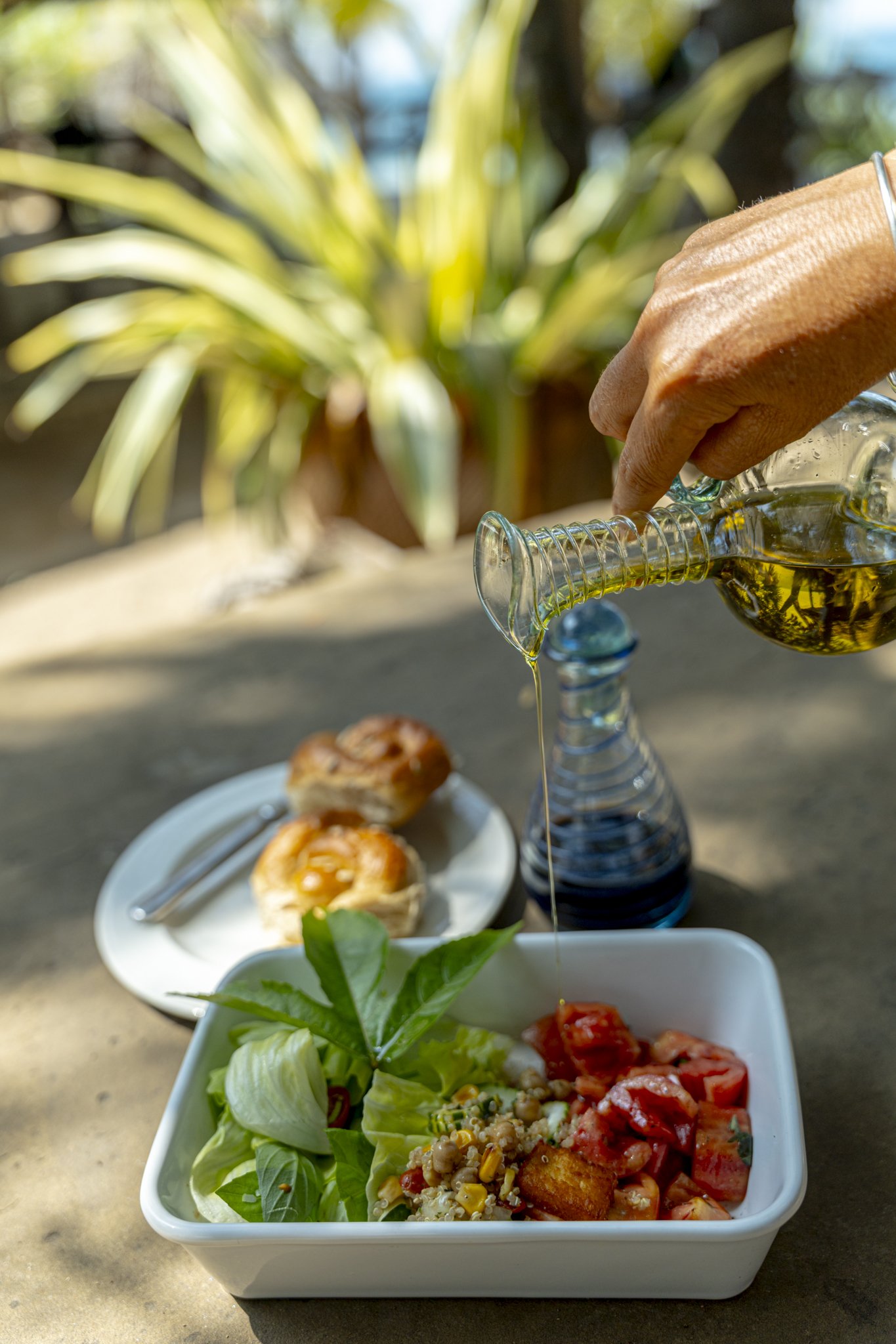 A bowl of salad with lettuce, tomato, and grilled chicken, topped with herbs and grains, on a table outdoors. Behind it, a plate of baked goods and a pitcher of dressing or oil being poured.