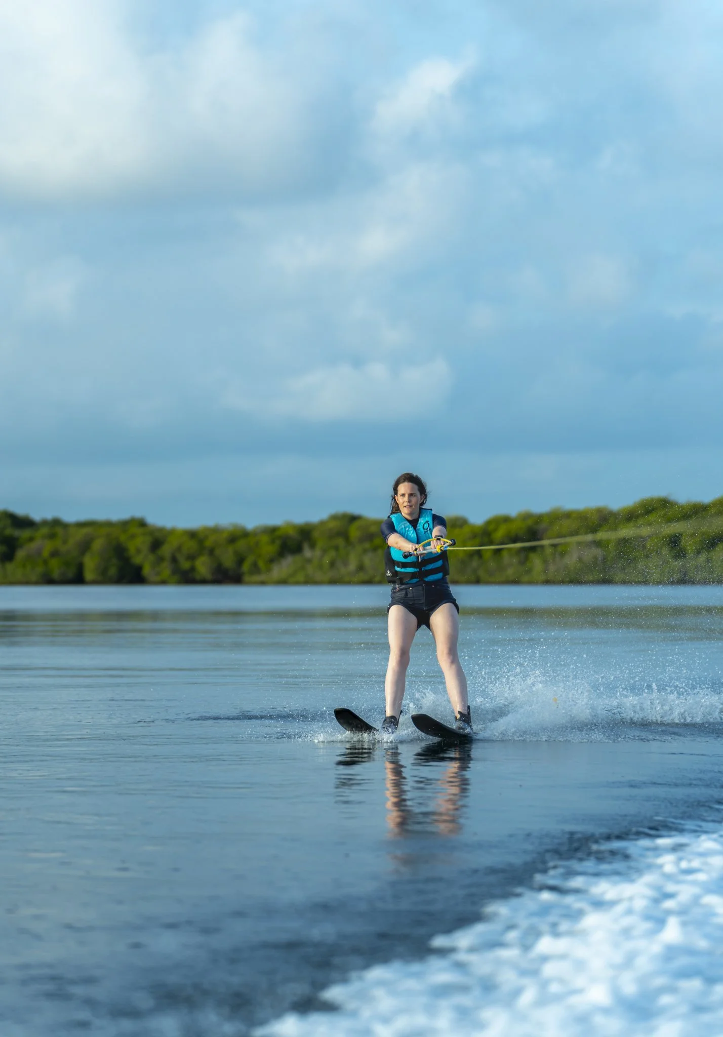 A woman wakeskates on a lake with greenery and a cloudy sky in the background.