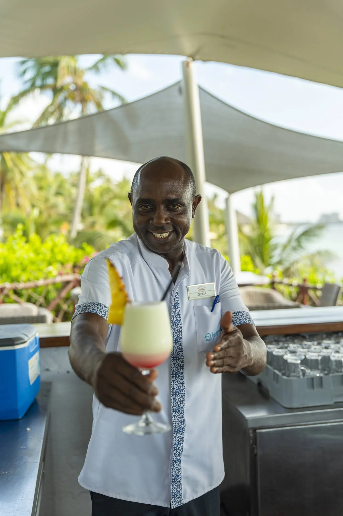 A man with a good smile, dressed in a white shirt with blue patterned trim, offering a layered cocktail drink in a tropical outdoor setting with green foliage and large sunshades.