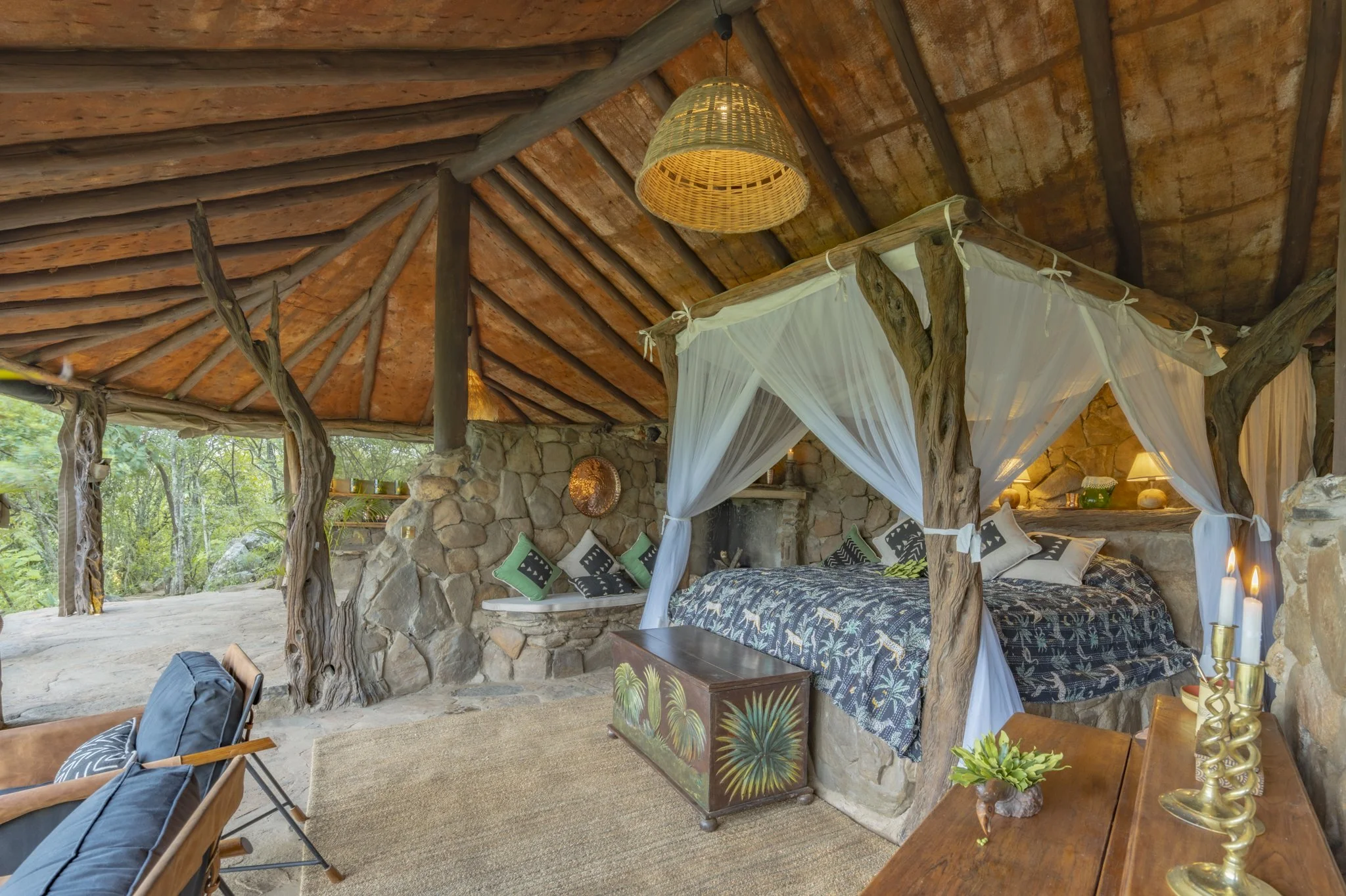 Interior of a rustic bedroom with a canopy bed featuring white drapes, a stone wall, wooden beams on the ceiling, and greenery outside visible through openings