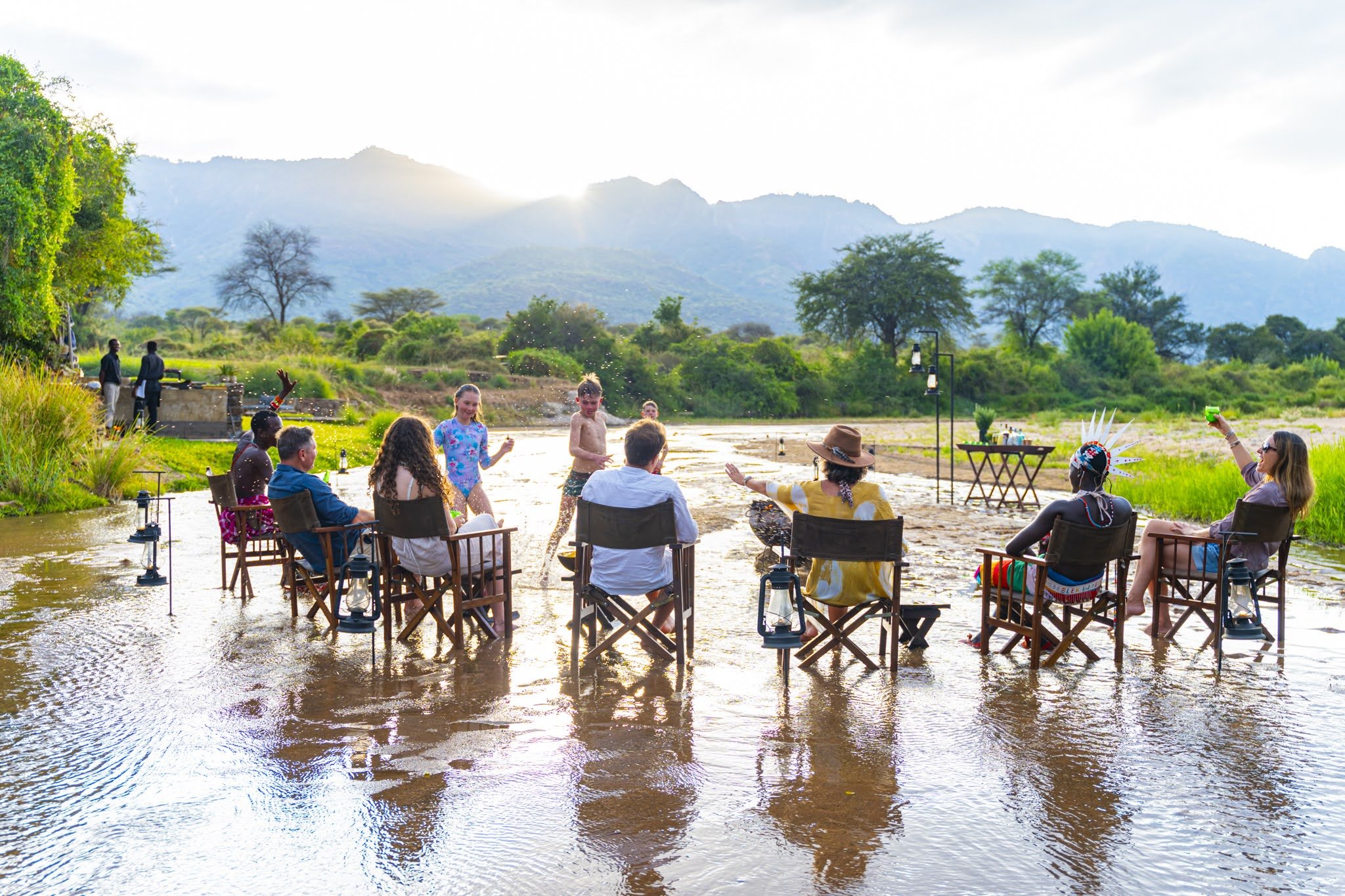 People sitting on chairs in a river with children playing and splashing water, surrounded by greenery and mountains in the background during daylight.