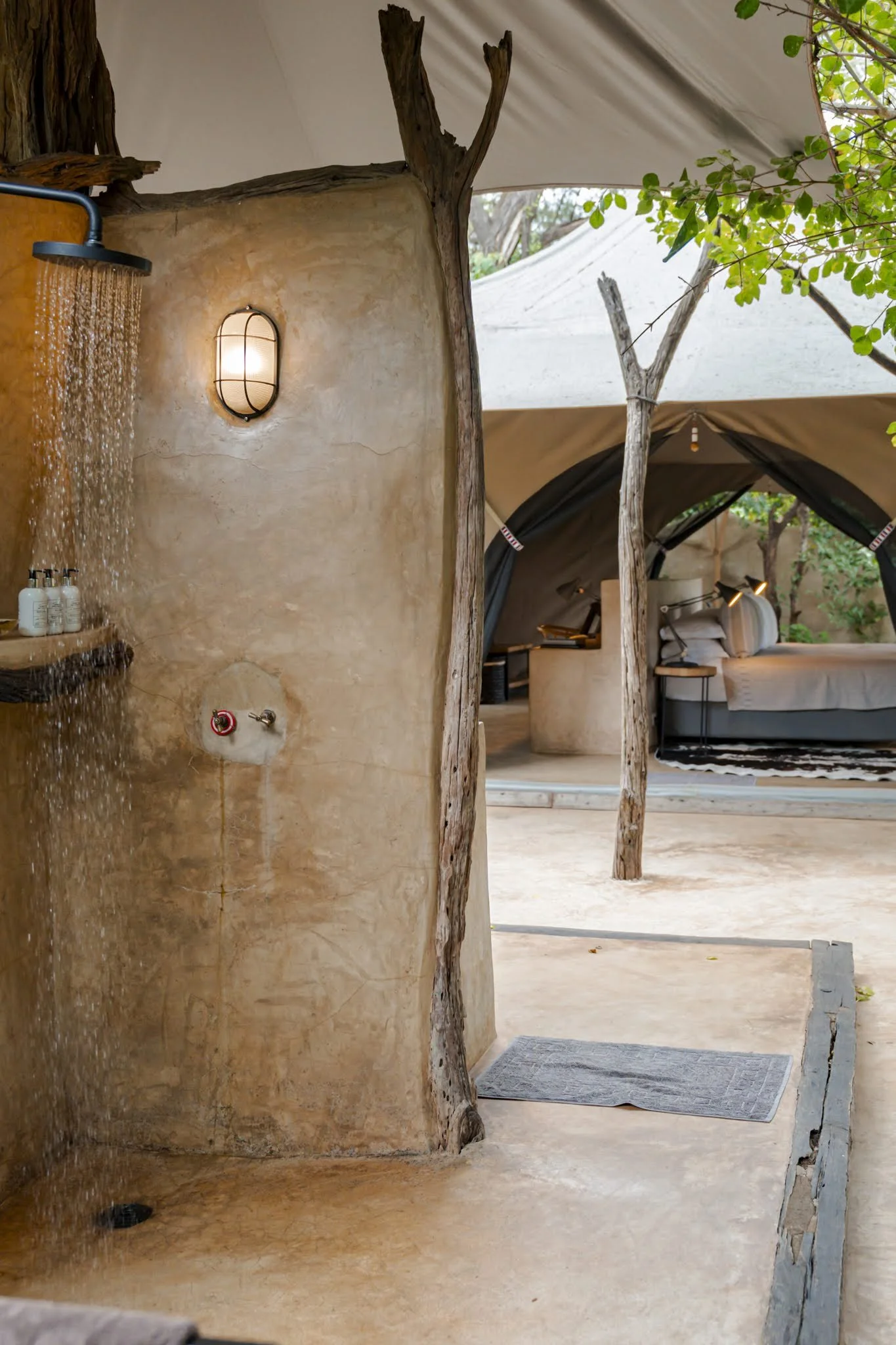 Outdoor shower area with a rain showerhead, streaming water onto a textured tan wall, with bottles of shampoo or soap on a shelf, nearby a bed inside a tent with beige and black fabric, and natural wooden supports, surrounded by greenery.