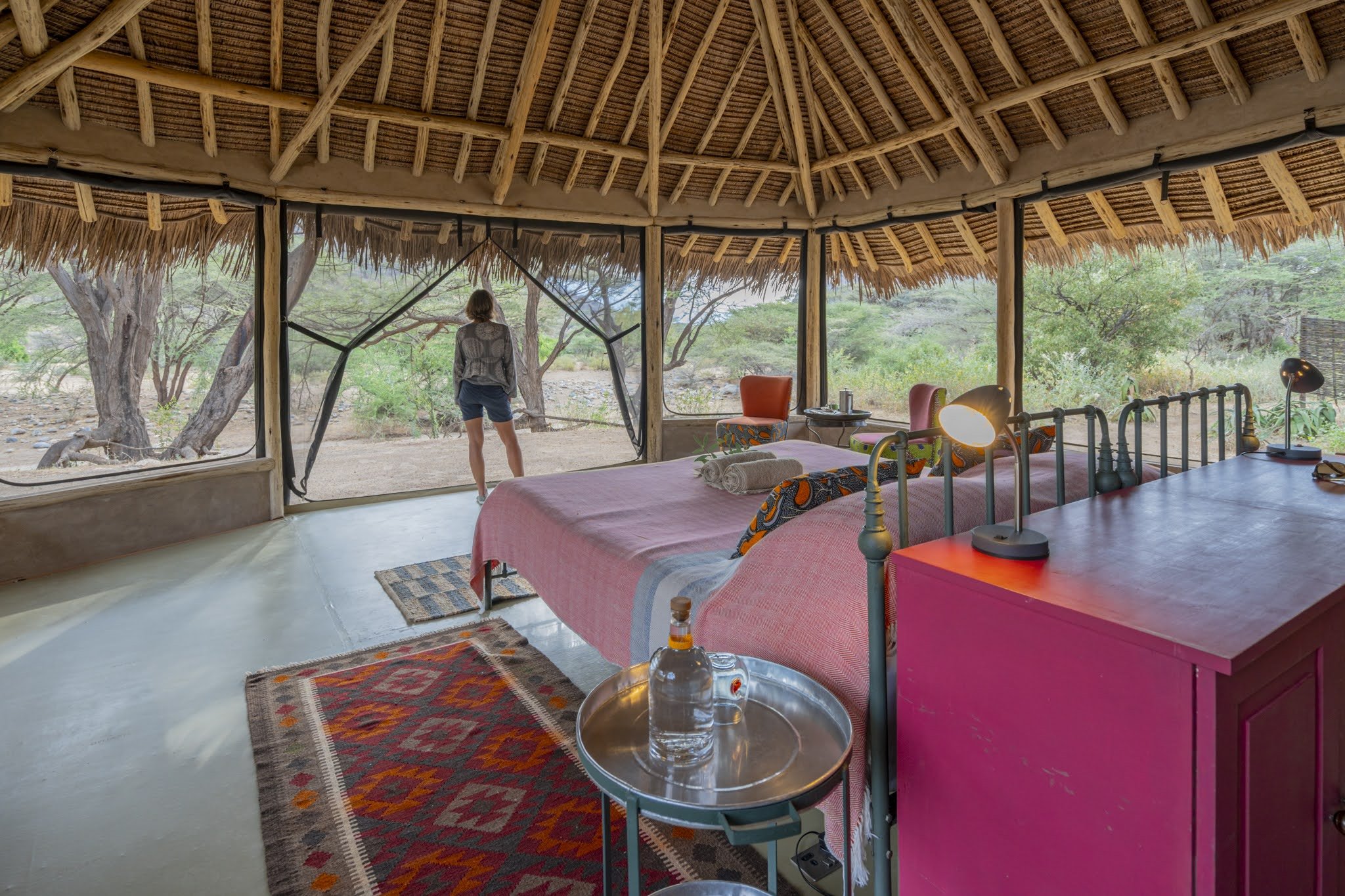 Interior of a thatched-roof safari lodge room with a bed, side table, and a large open window showing trees outside, with a woman standing and looking out.