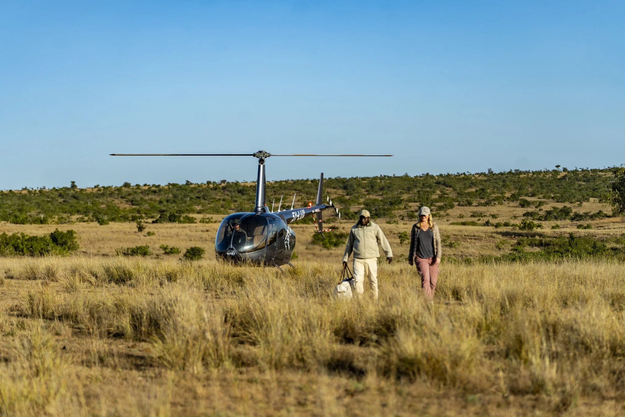 A helicopter on a grassy field with two people walking nearby, set against a backdrop of rolling hills and a clear blue sky.