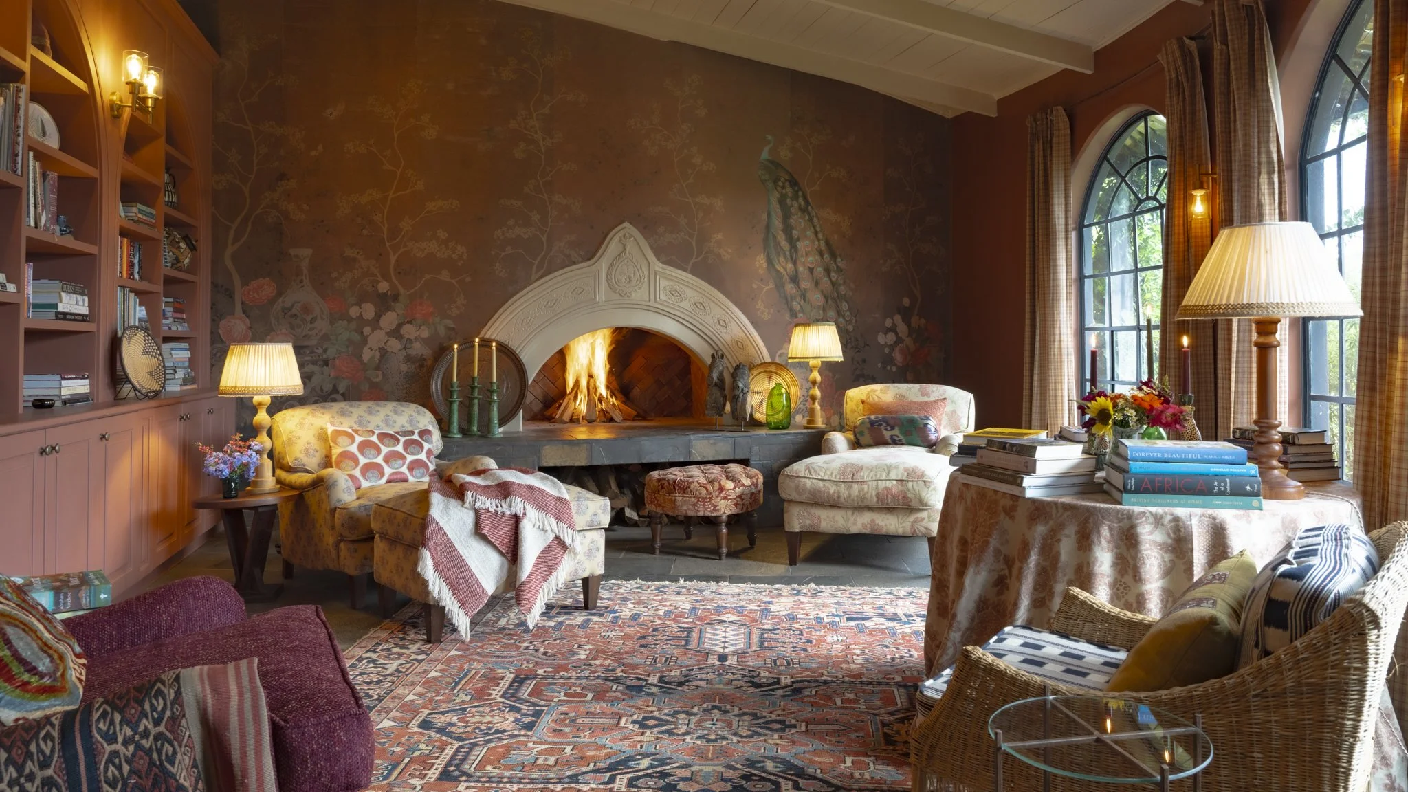 A cozy living room with a fireplace, floral wallpaper featuring a peacock, and large arched windows with plaid curtains. The room has multiple with lamps, books, flowers, and patterned furniture, creating a warm, inviting atmosphere.