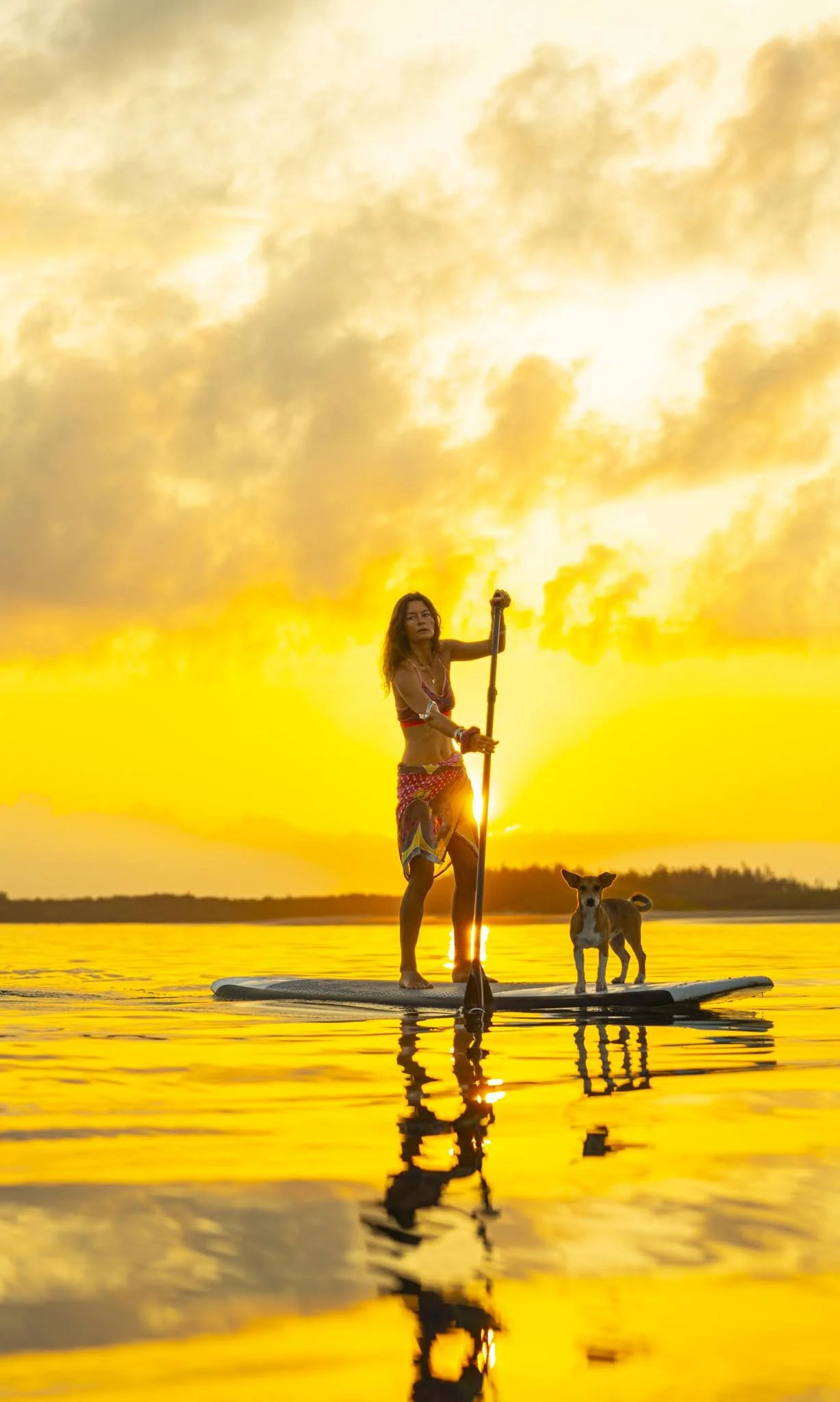 Woman paddleboarding with a dog at sunset on a lake.