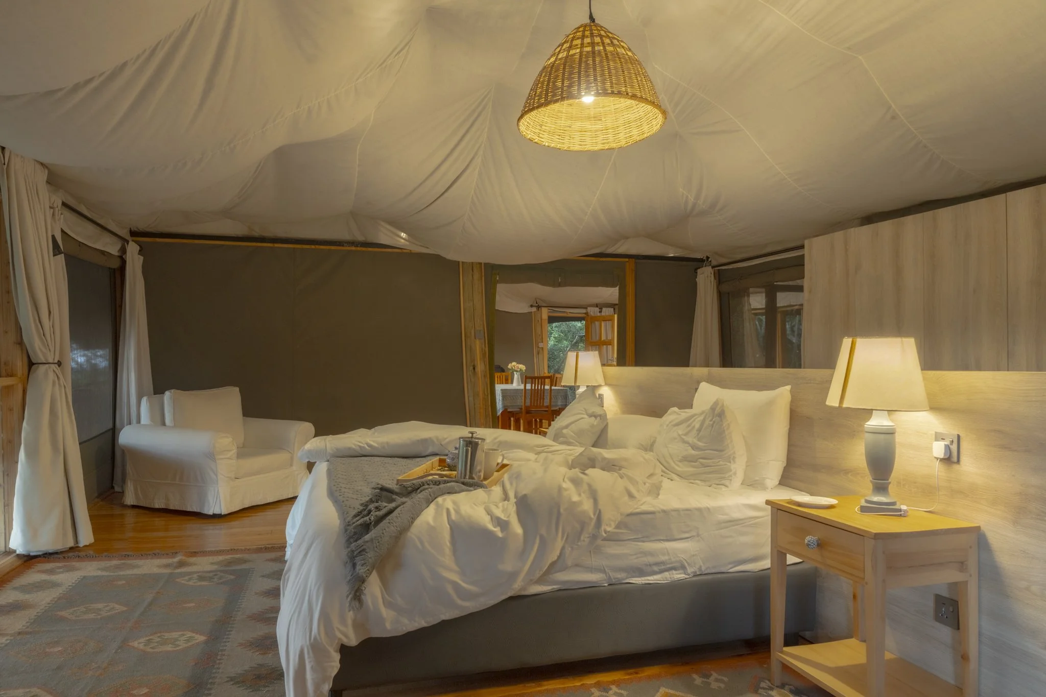 A rustic bedroom with a large bed, white bedding, and a wooden headboard. There is a nightstand with a white lamp beside the bed. A wicker lampshade hangs from the ceiling, and the ceiling is covered with a white fabric. The room has windows with white curtains, and a small sitting area with a white armchair and a wooden dining table with chairs in the background. The floor is wooden, and a patterned area rug is partially visible.