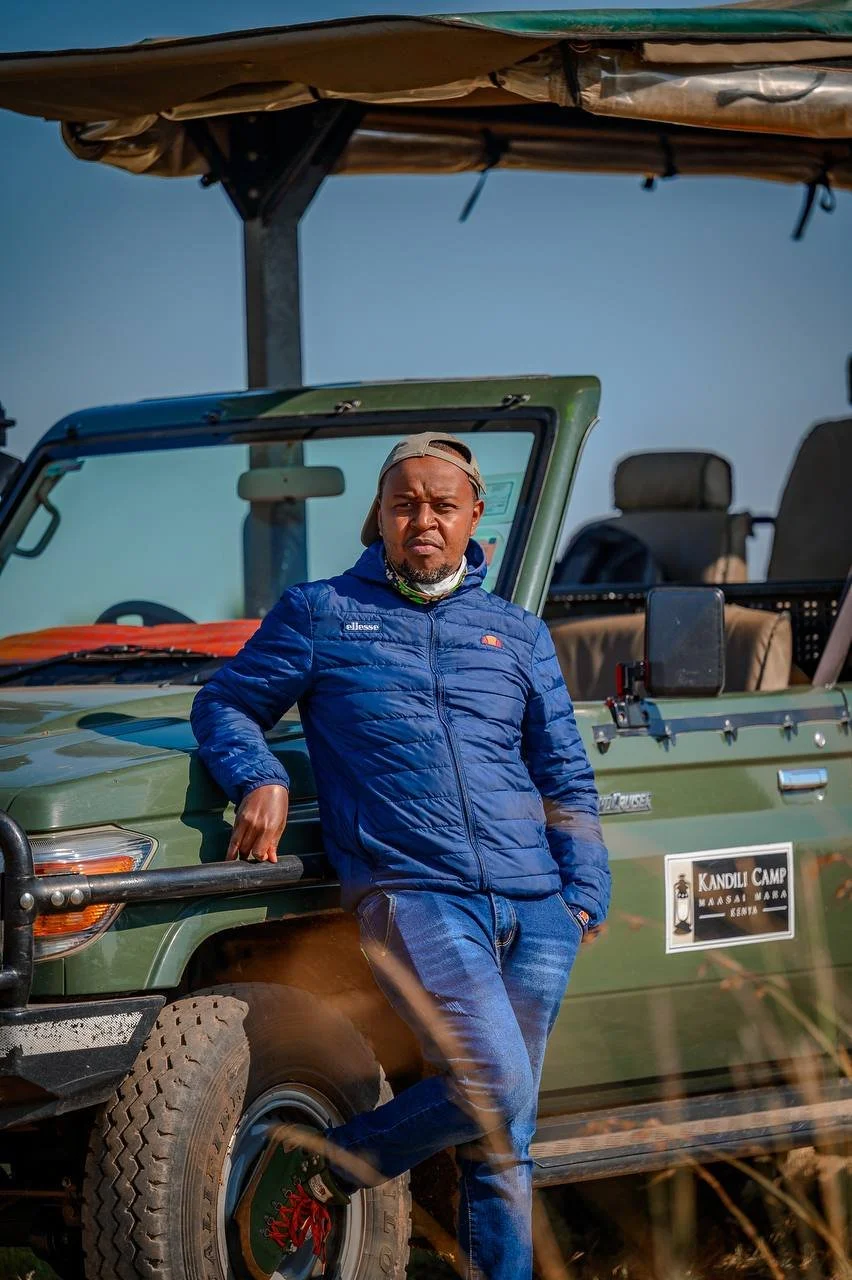 A man leaning against a green off-road vehicle with a canopy, wearing a blue jacket, jeans, and a backwards baseball cap, in a rural outdoor setting with a clear blue sky.