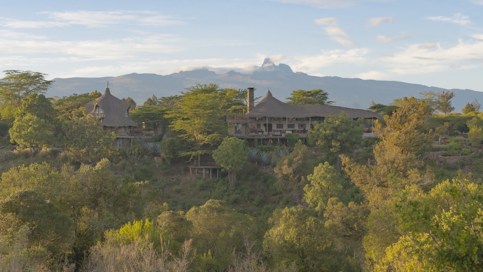 Scenic view of lush green trees, traditional thatched-roof houses on a hillside, with a mountain in the background under a partly cloudy sky.