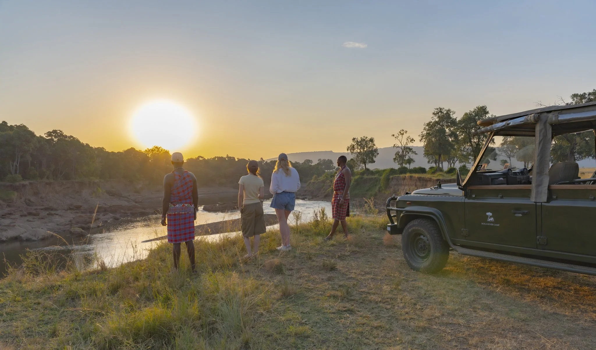 A dramatic sunset at the Mara River overlooking the Ololo Escarpment with guest enjoying a sundowner with their Maasai safari guides and a Land Rover besides them.