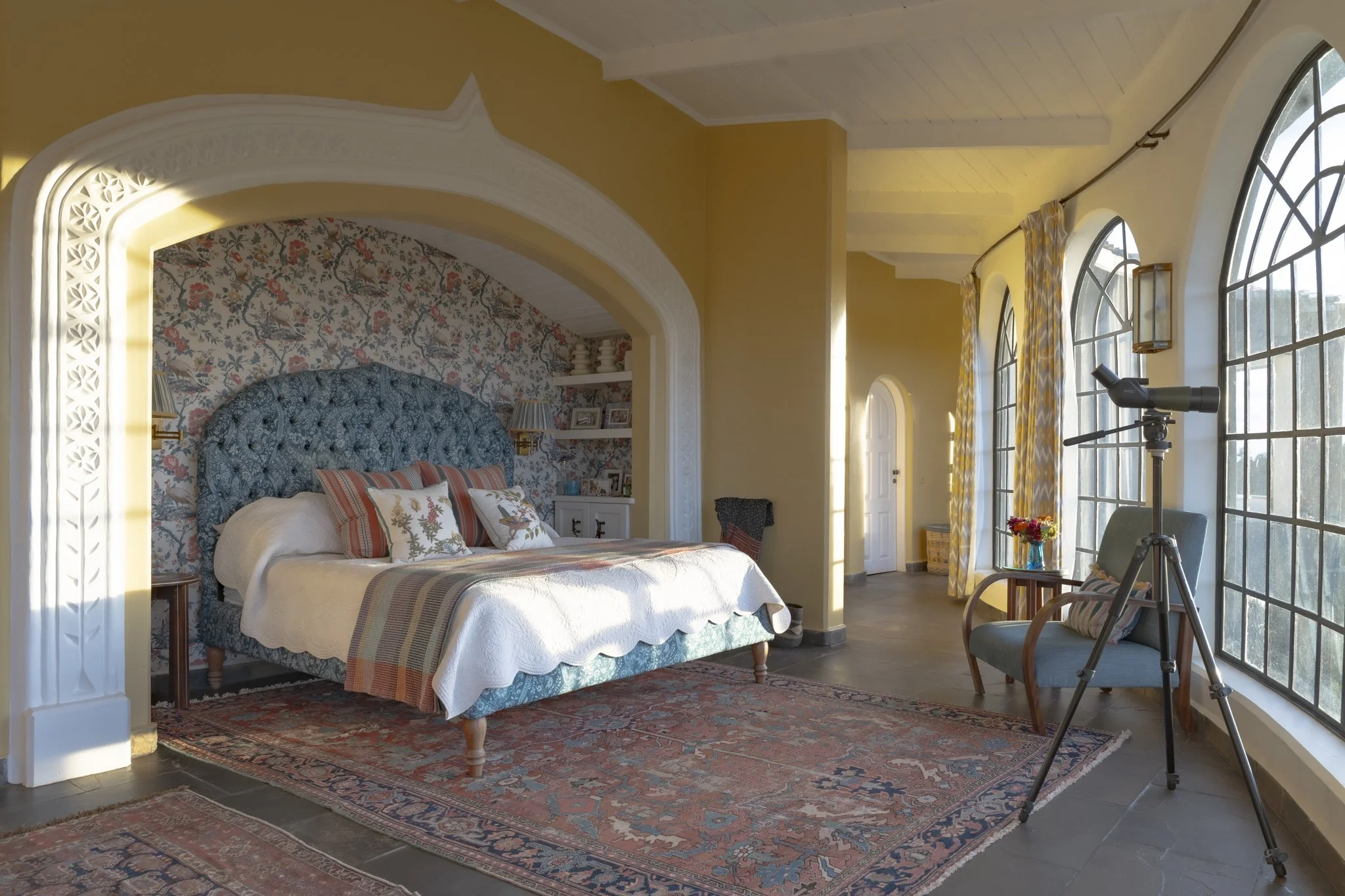 A bedroom with a large bed, upholstered headboard, floral wallpaper behind the bed, and decorative pillows. The room has three arched windows with patterned curtains, a telescope nearby, and a colorful rug on the floor.