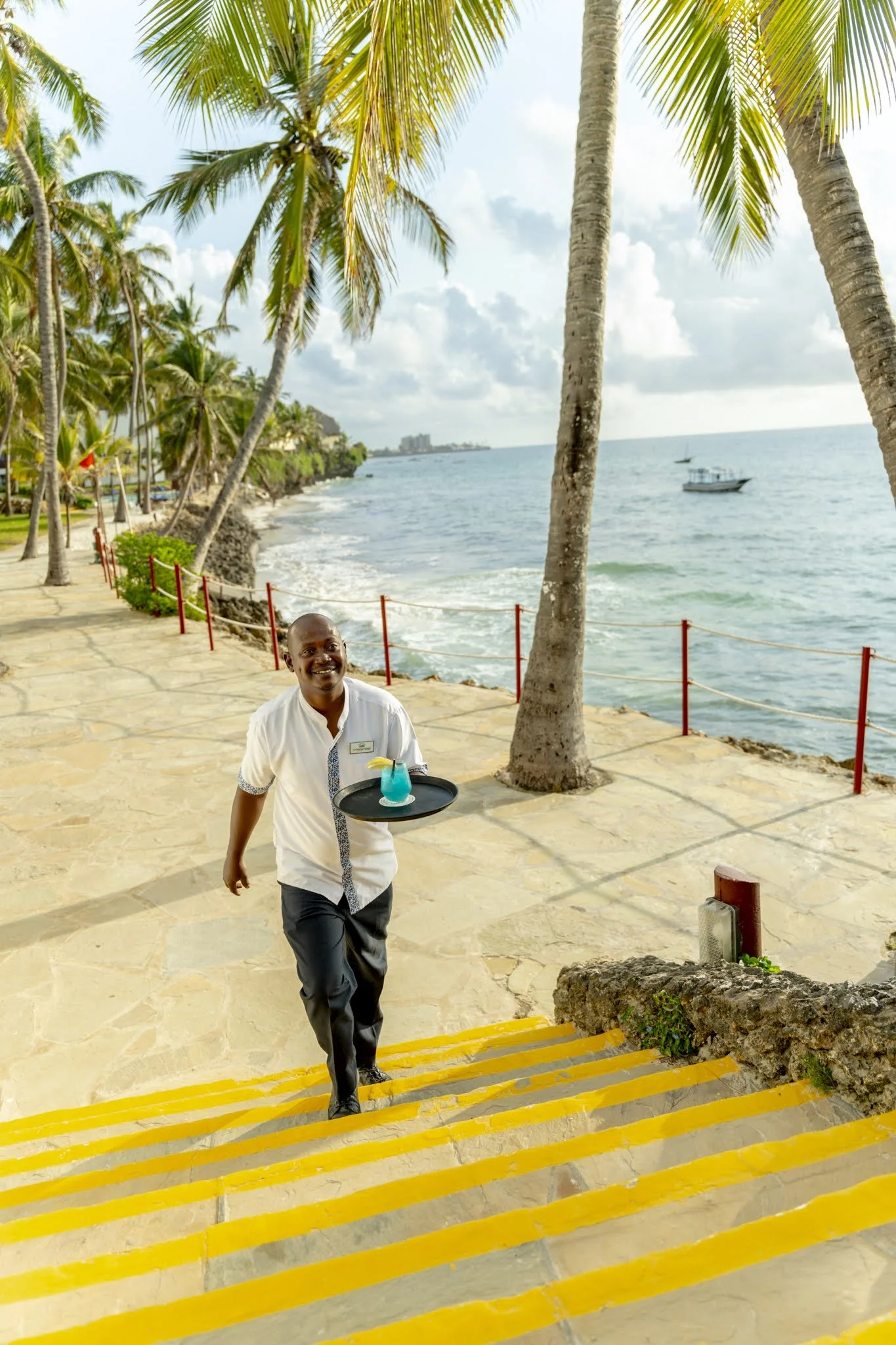 A smiling waiter walking up outdoor yellow striped stairs beside a sandy pathway near the ocean, with tall palm trees and boats in the background on a sunny day.