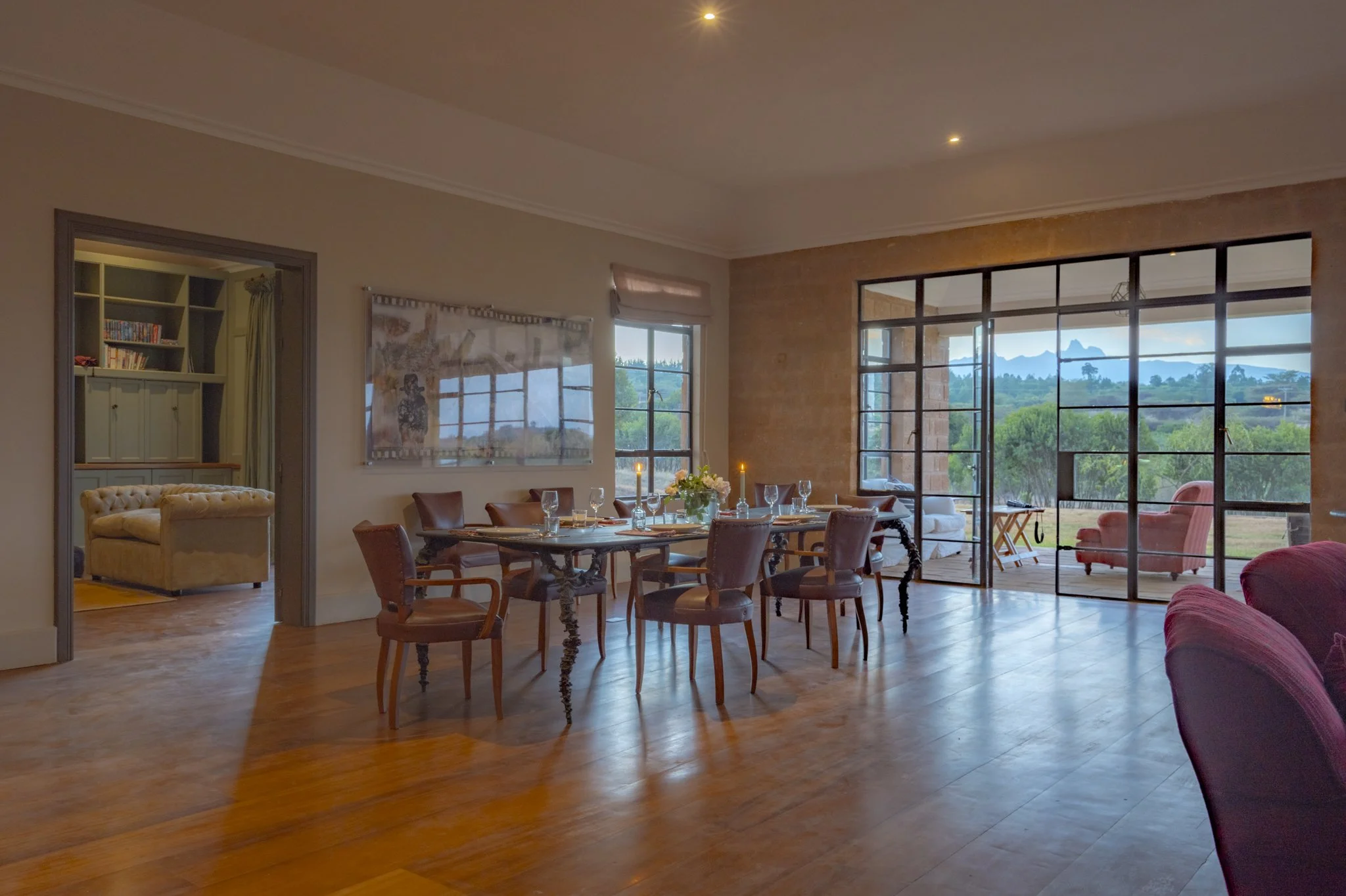 Living room with large windows and view of greenery and mountains. Dining table set with candles and flowers, adjacent to a cozy seating area with chairs and sofas.