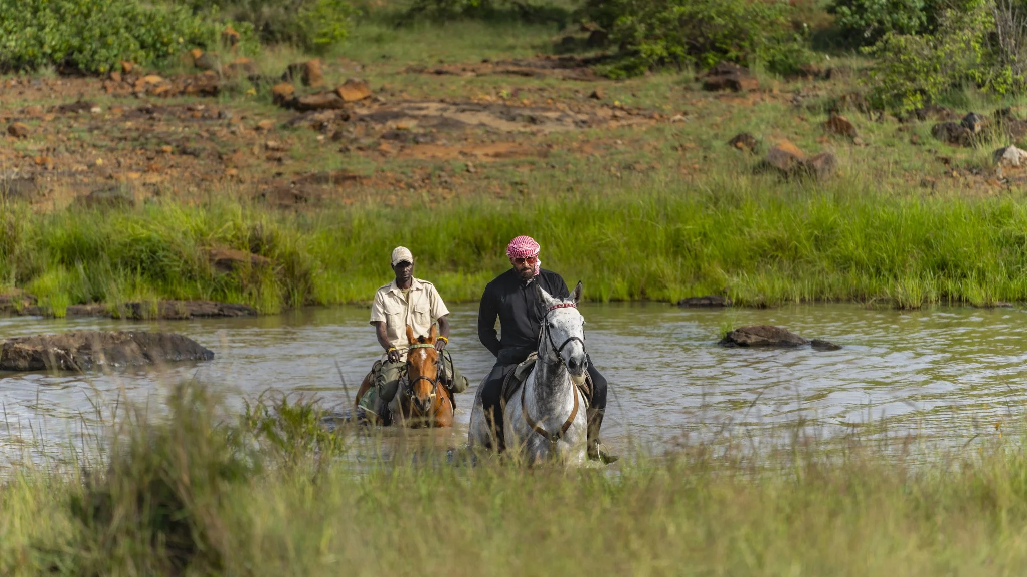 Two men riding horses through a river with green grass and shrubs in the background.