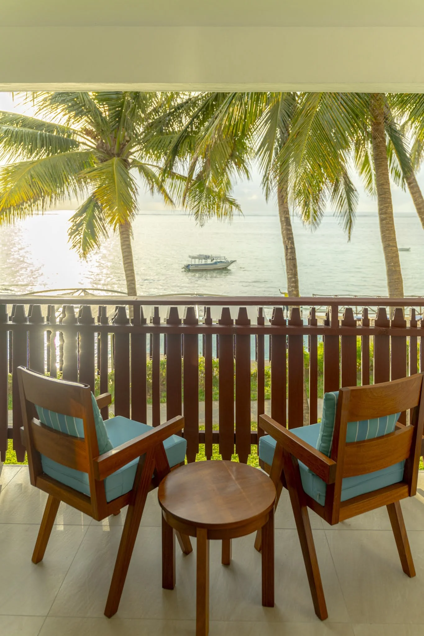 Balcony view with two wooden chairs with blue cushions and pillows, overlooking palm trees, water, and a boat in the distance.