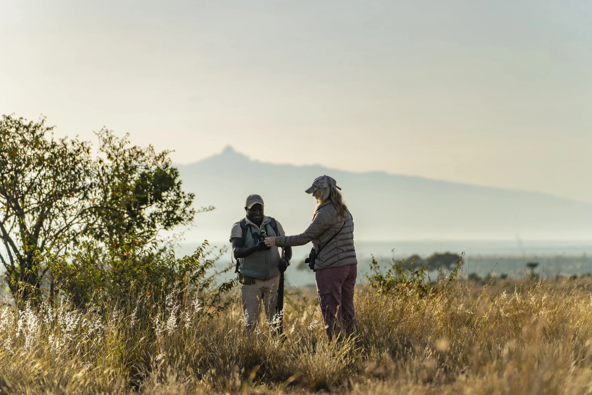 Two people are standing outdoors in a field with tall grass and bushes, exchanging something. They are wearing outdoor gear including hats, and there is a scenic background with mountains and a clear sky.