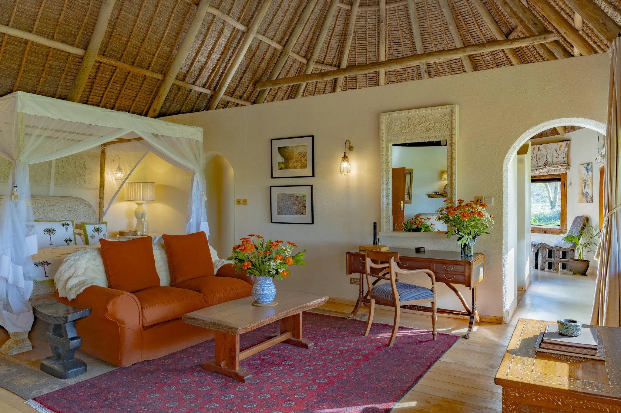 Beautiful bedroom with a huge bed with a net plus an orange sofa with flowers on the table and desk with photo prints adorning the walls and a thatched roof in place.