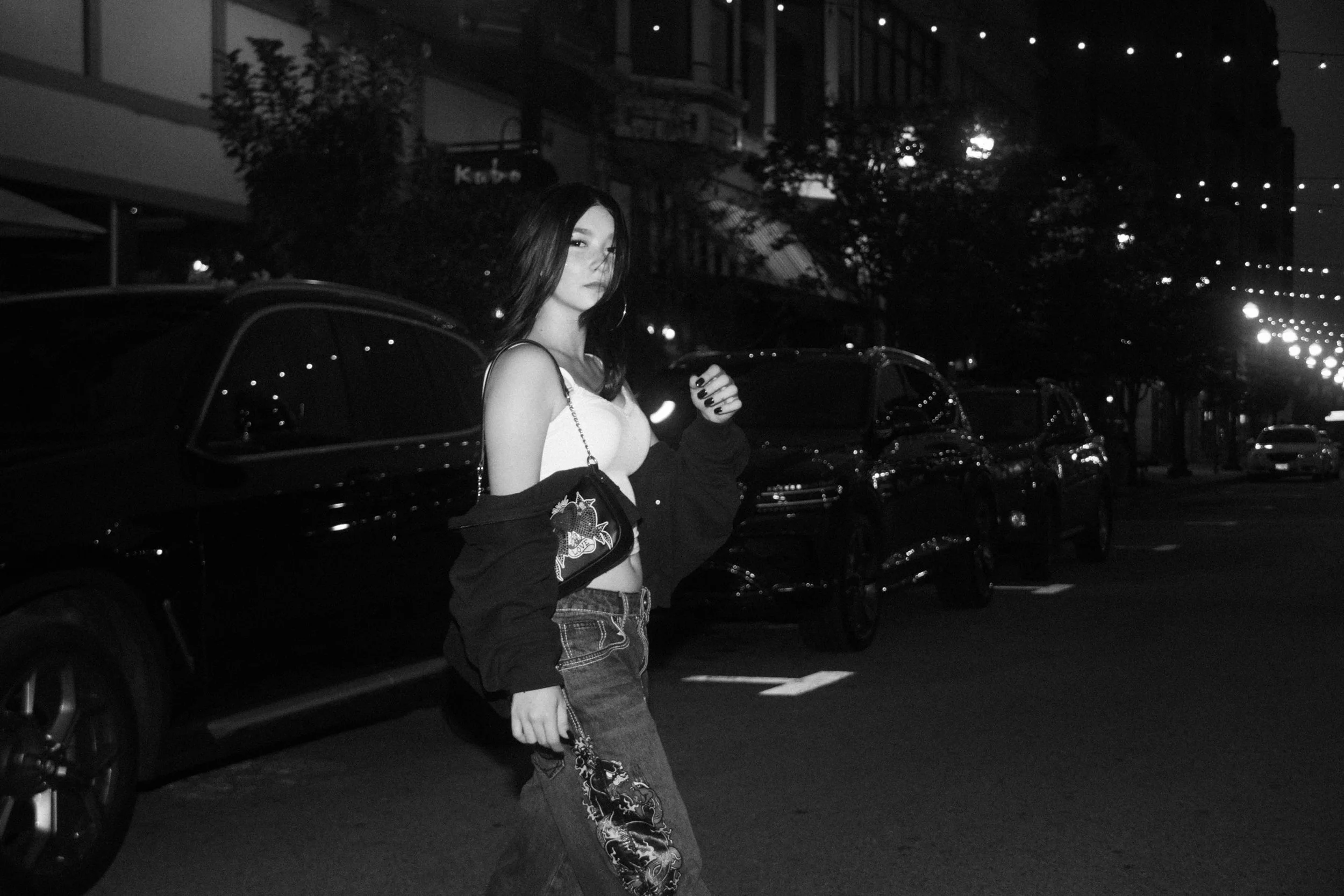 A woman with long dark hair, wearing a white sleeveless top, large hoop earrings, and patterned jeans, stands beside a dark car on a city street at night. The background shows buildings with lit windows and string lights overhead.