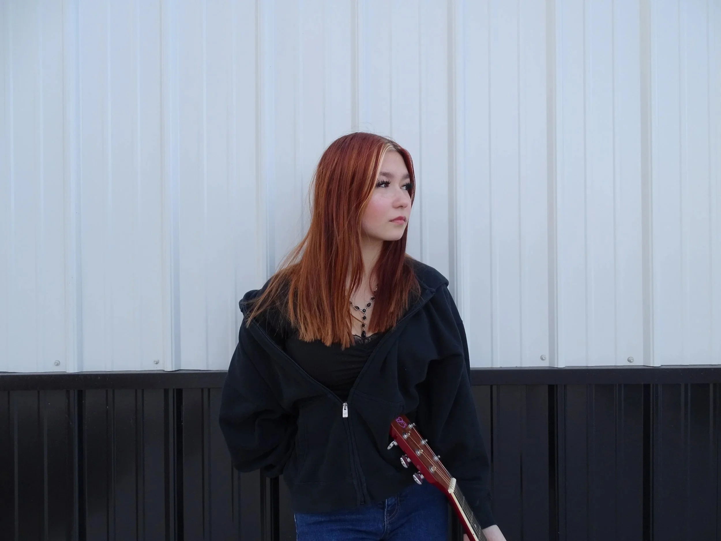 A young woman with long red hair standing against a light-colored, vertically paneled wall, holding a red electric guitar.