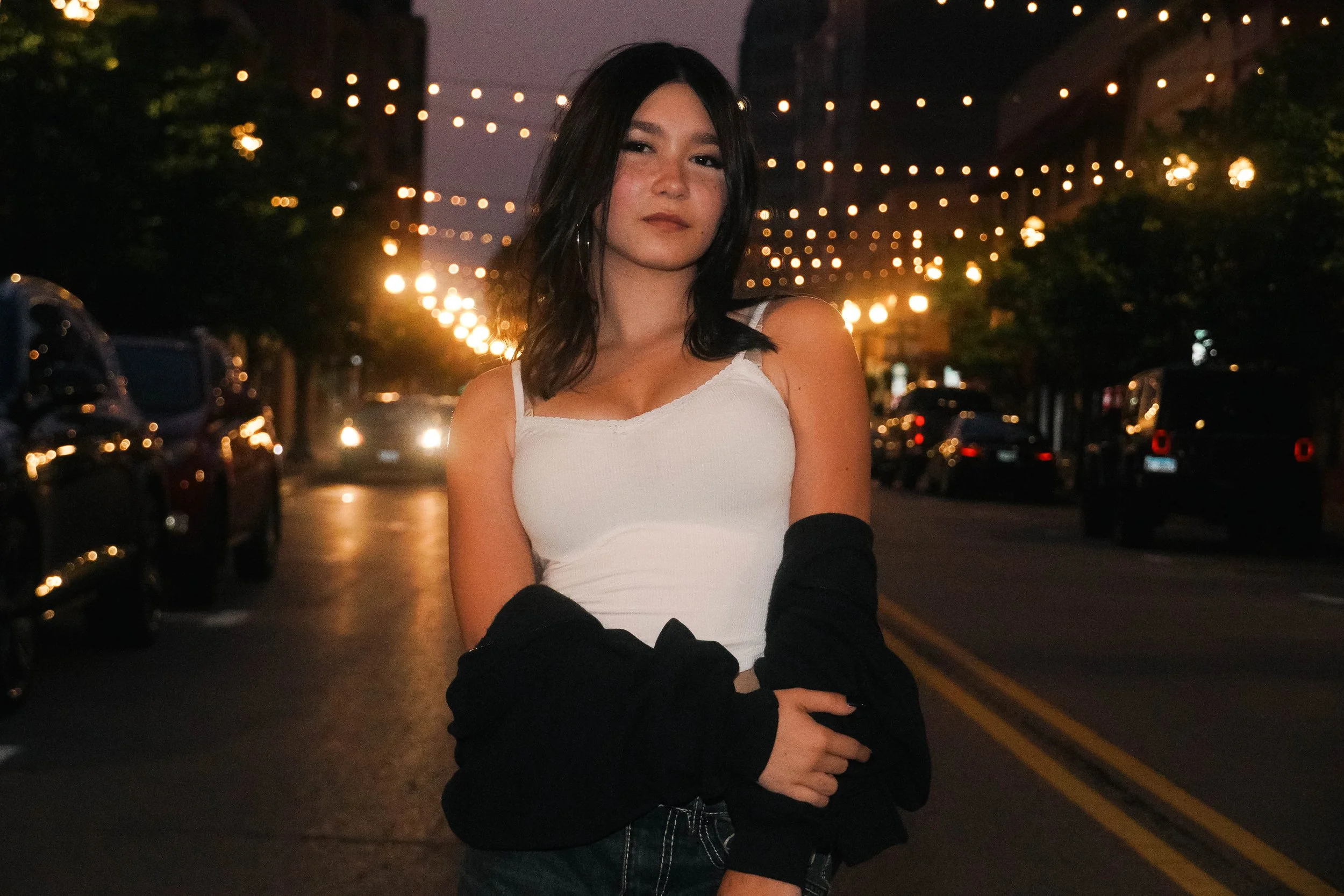 A young woman with black hair and fair skin standing on a city street at night, illuminated by string lights overhead, wearing a white tank top and black arm sleeves. Cars are parked along the street, and the background shows buildings and trees.