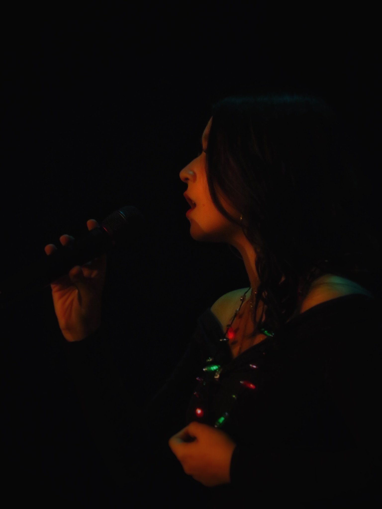 A woman singing into a microphone while holding a phone in her hand, wearing headphones and a necklace with colored lights.
