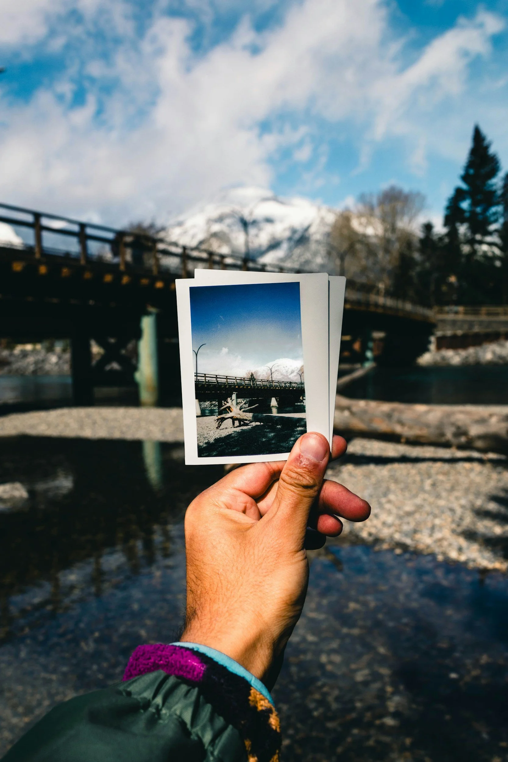 Person holding a printed photo of a bridge over a river with mountains and a cloudy sky in the background.