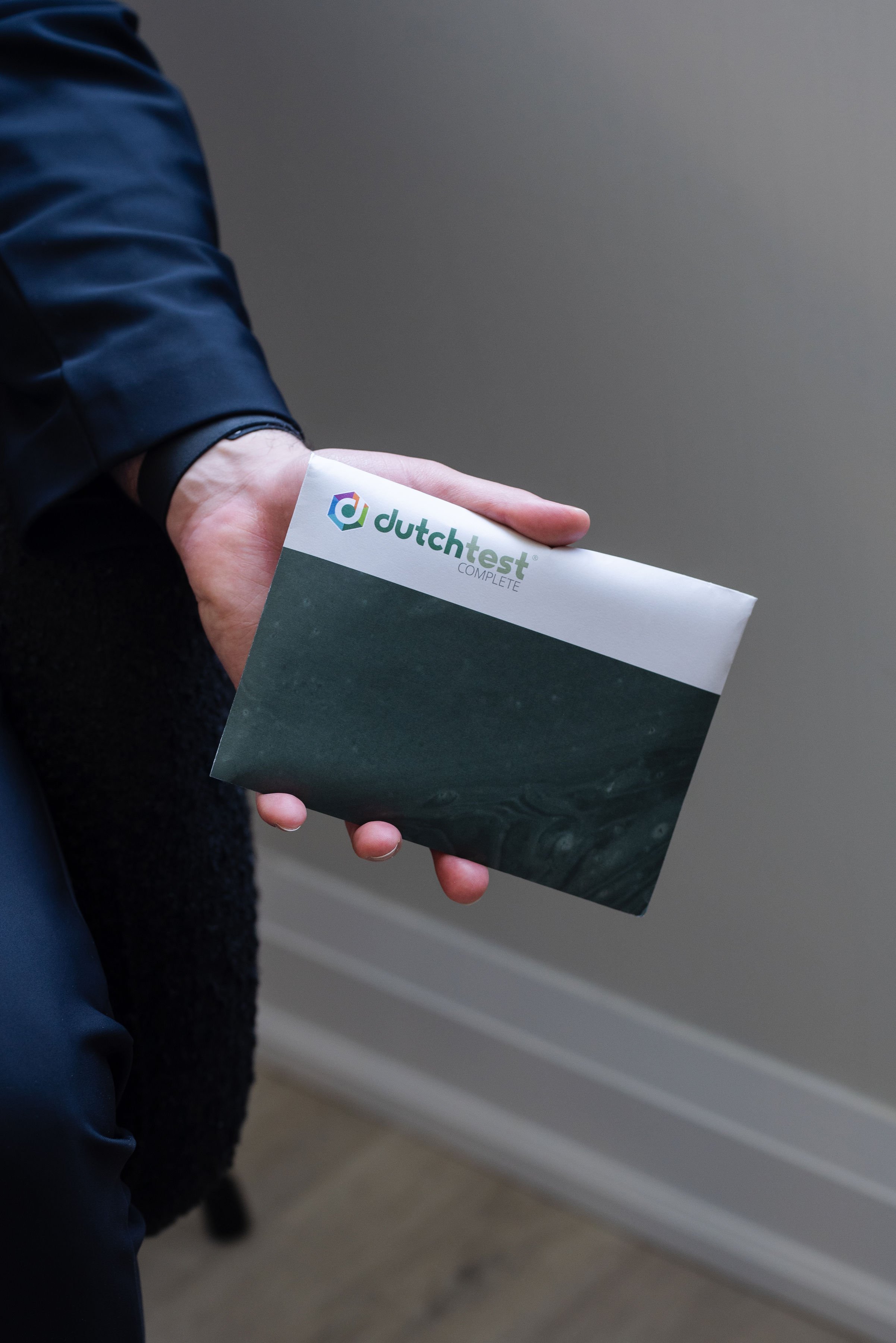 A person wearing a dark jacket holding a booklet titled 'dutch test COMPLETE' in front of a gray background.