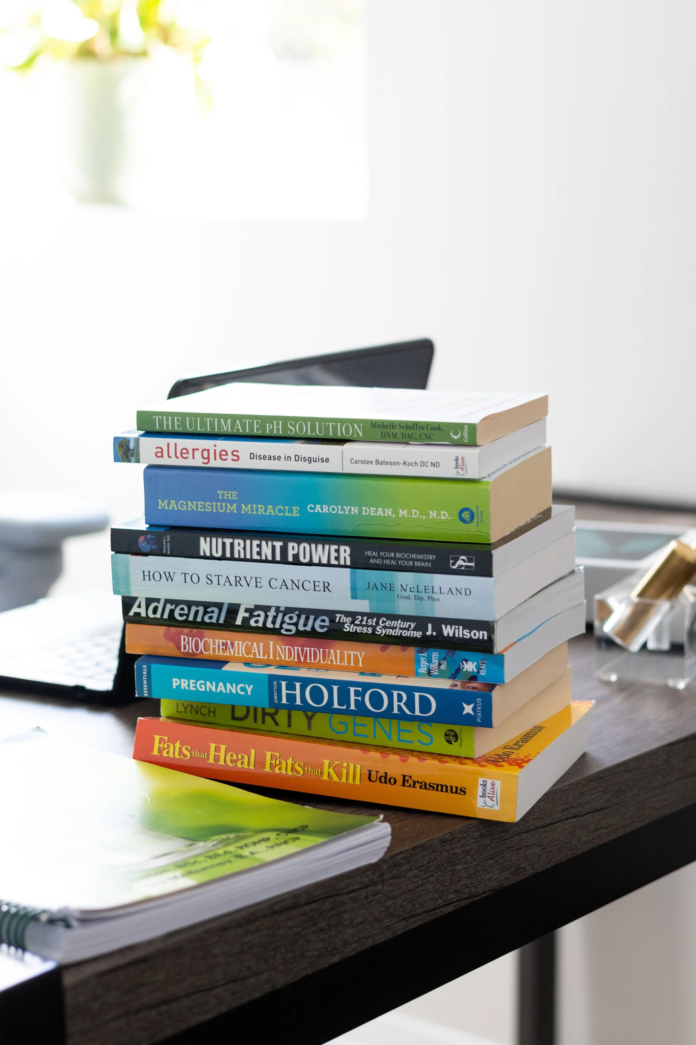 A stack of health and science books on a wooden desk in a well-lit room, with a potted plant in the background.