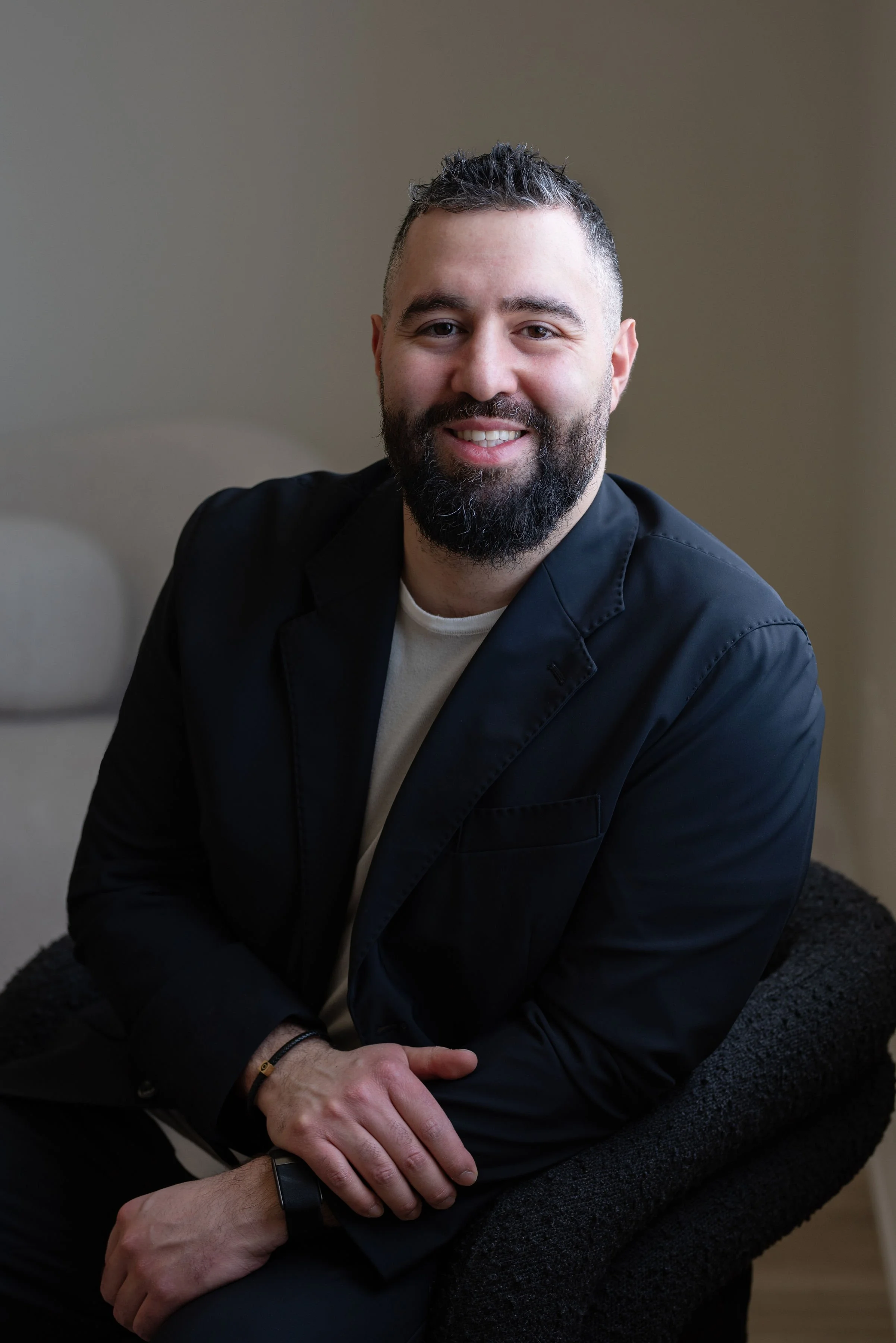 Portrait of a smiling man with dark, short spiked hair and a beard, dressed in a black jacket and sitting on a chair with a neutral background.