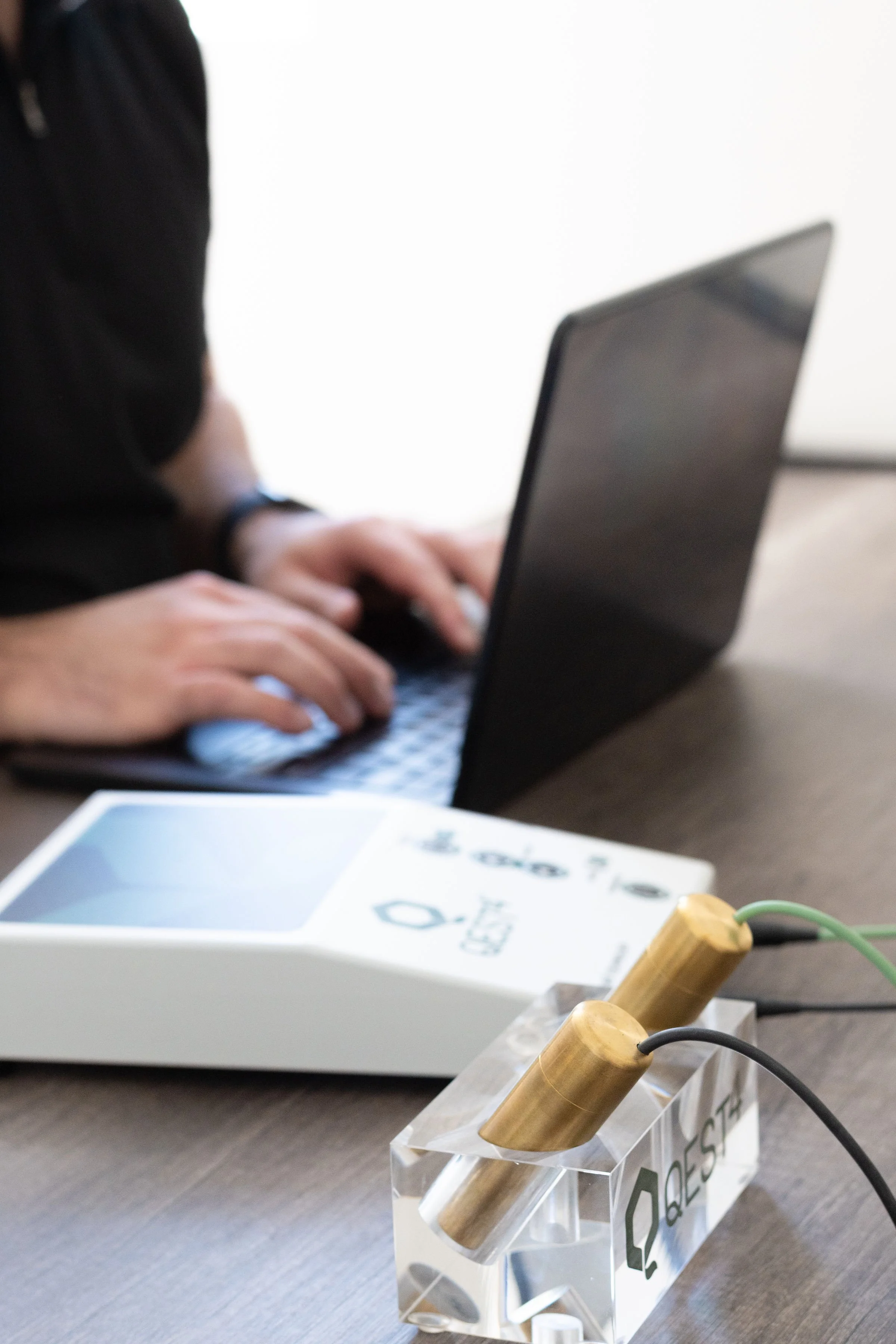 Close-up of electrical wires and a clear plastic container with a logo, holding two wooden electrical plug casings, on a grey table in an office or workshop setting.
