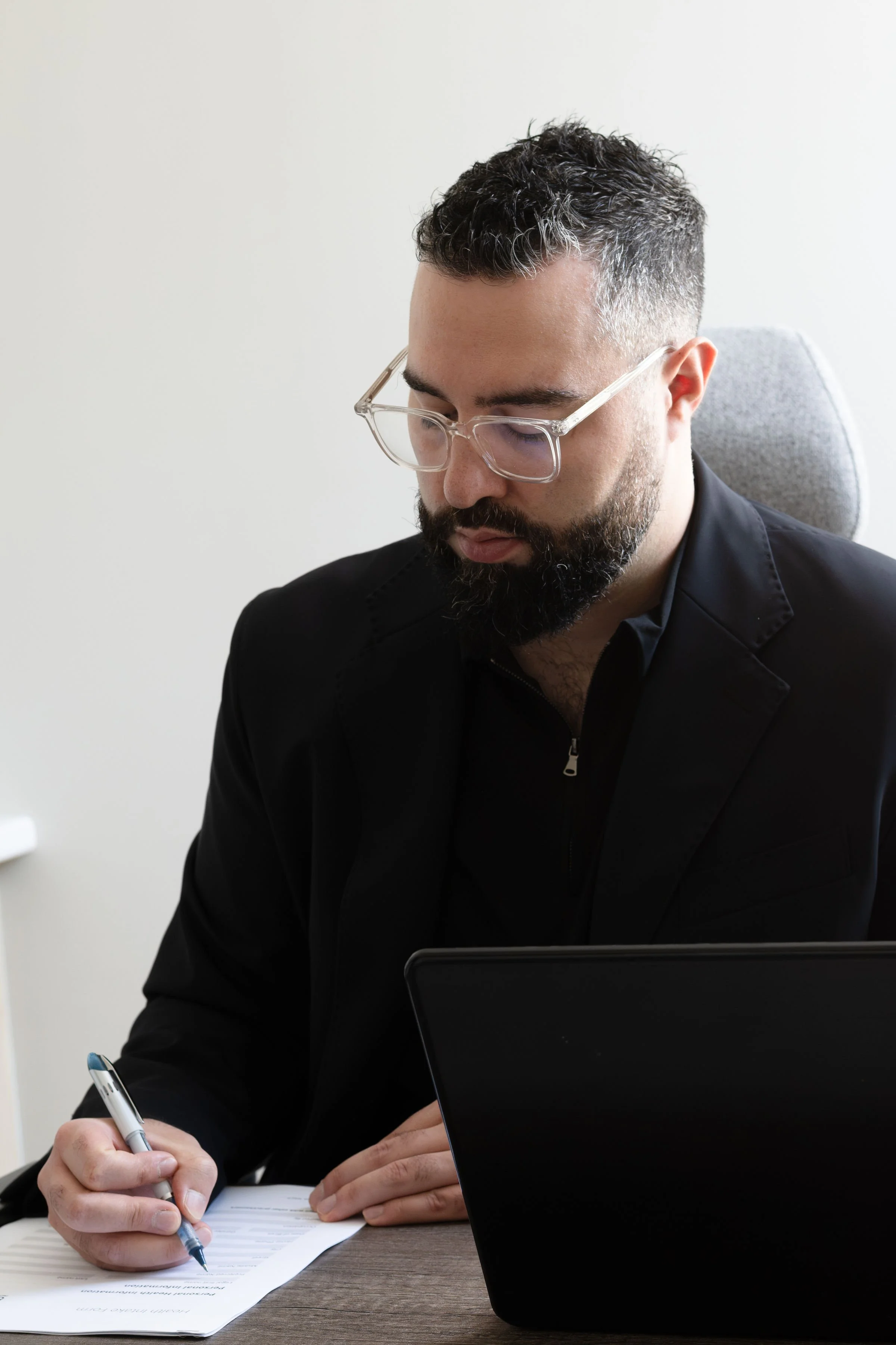 A man with glasses, a beard, wearing a black jacket, sitting at a desk, writing on a piece of paper with a pen, with a laptop in front of him and a neutral background.